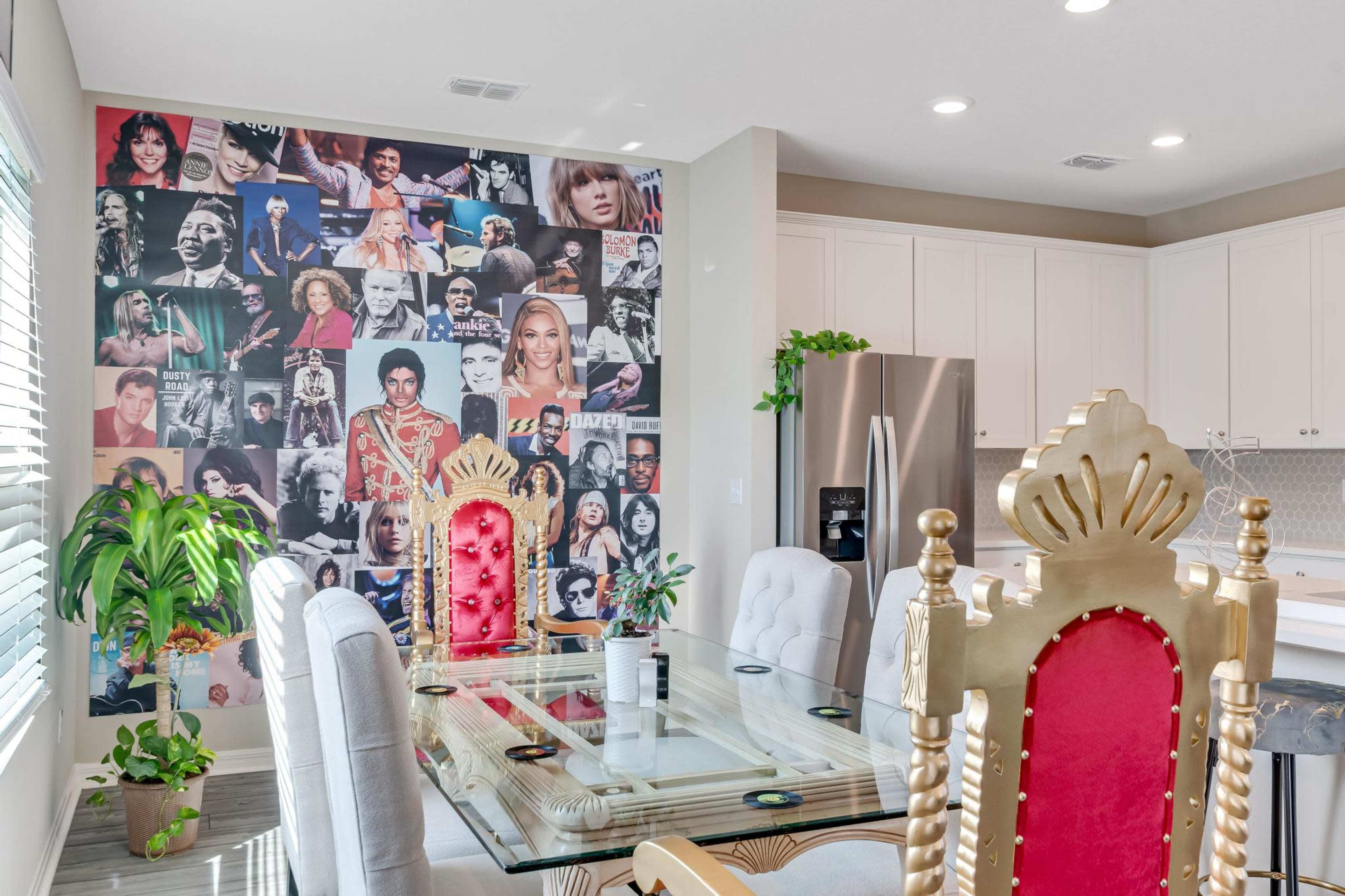 A dining area with a glass table and ornate golden chairs, featuring a wall collage of various celebrities.