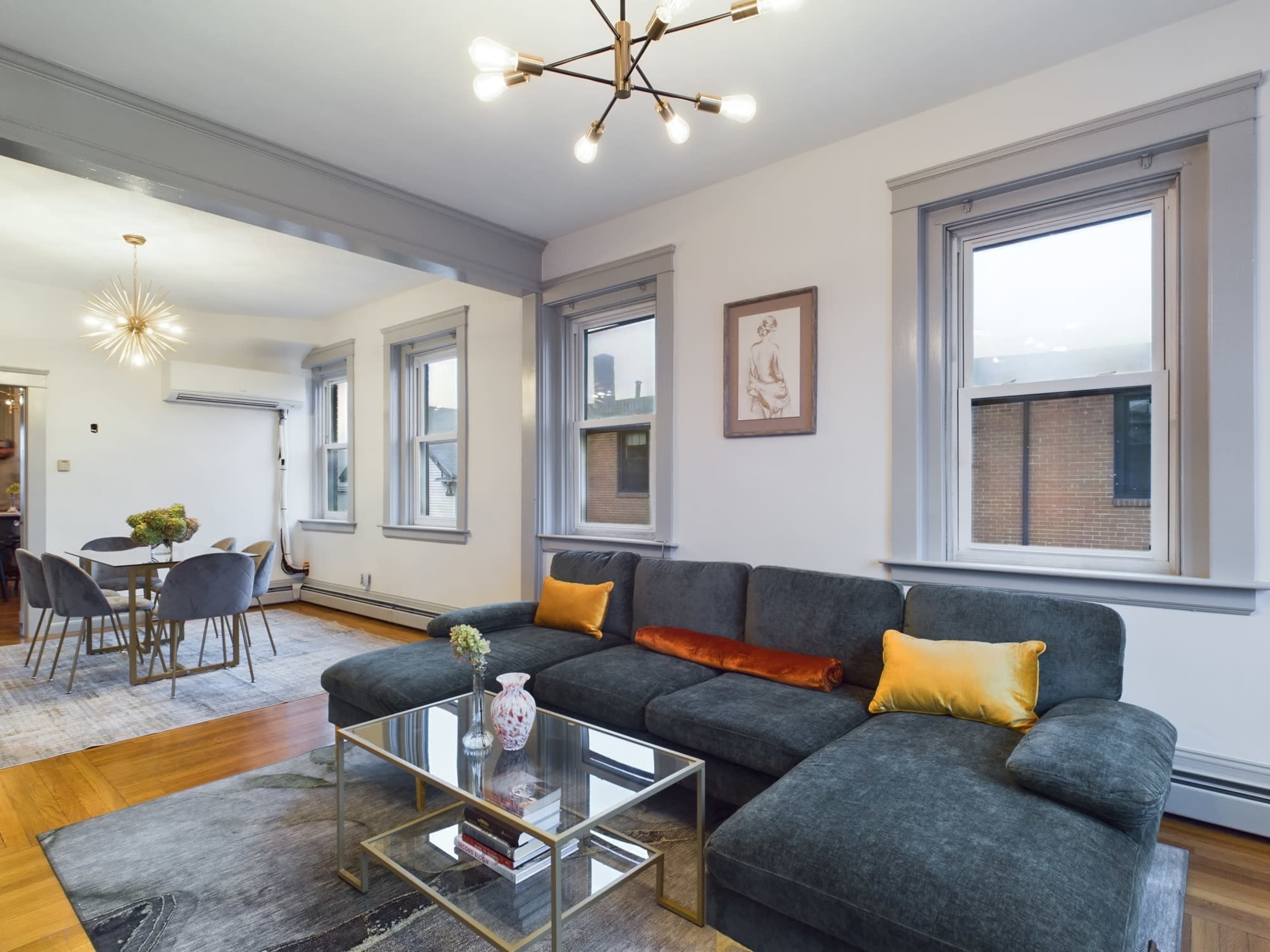 A modern living room with a dark gray sectional sofa, a glass coffee table, and two large windows, leading to a dining area with a round table and chairs.