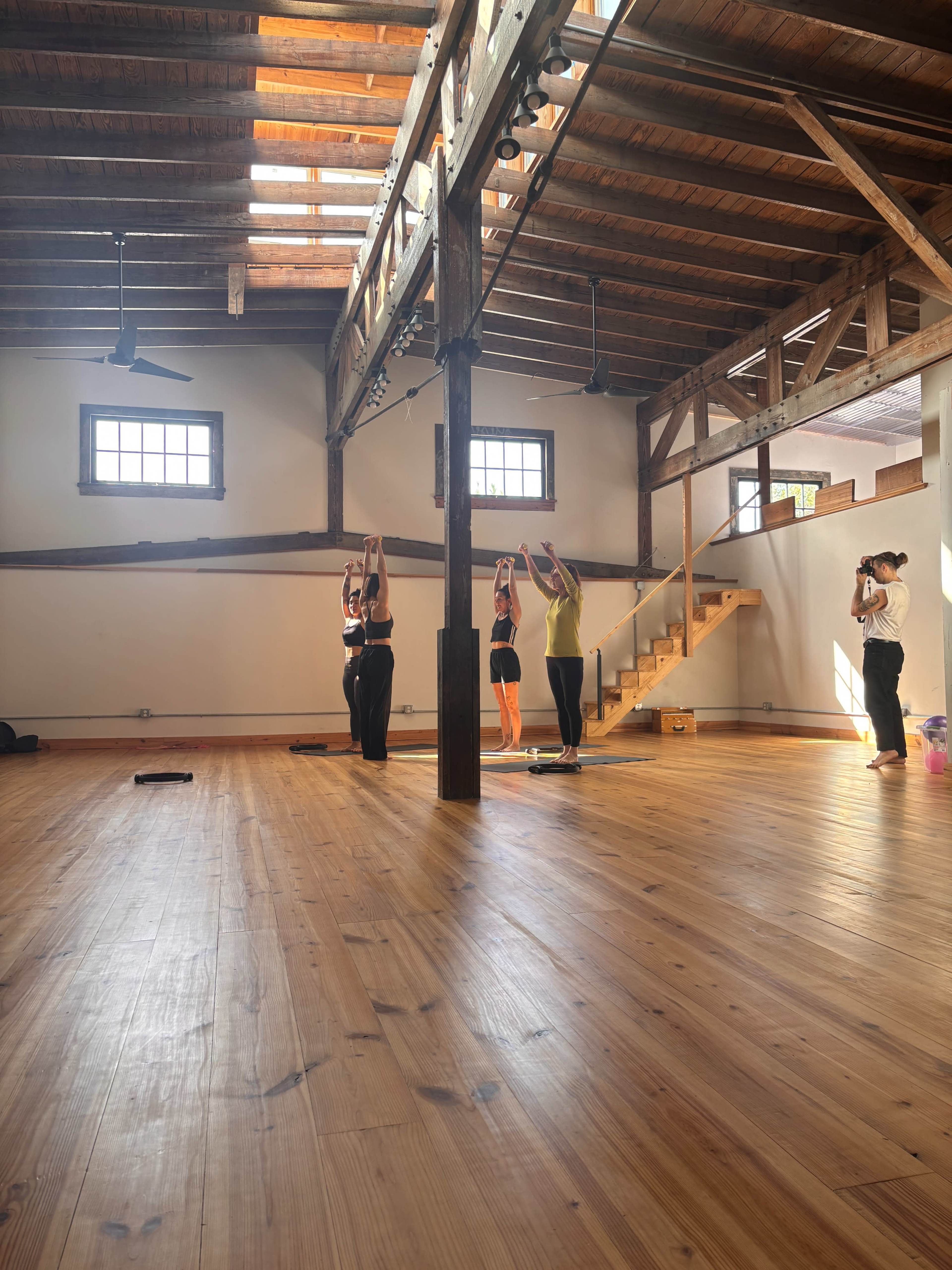 Four individuals are participating in a yoga session in a spacious room with wooden beams and large windows.