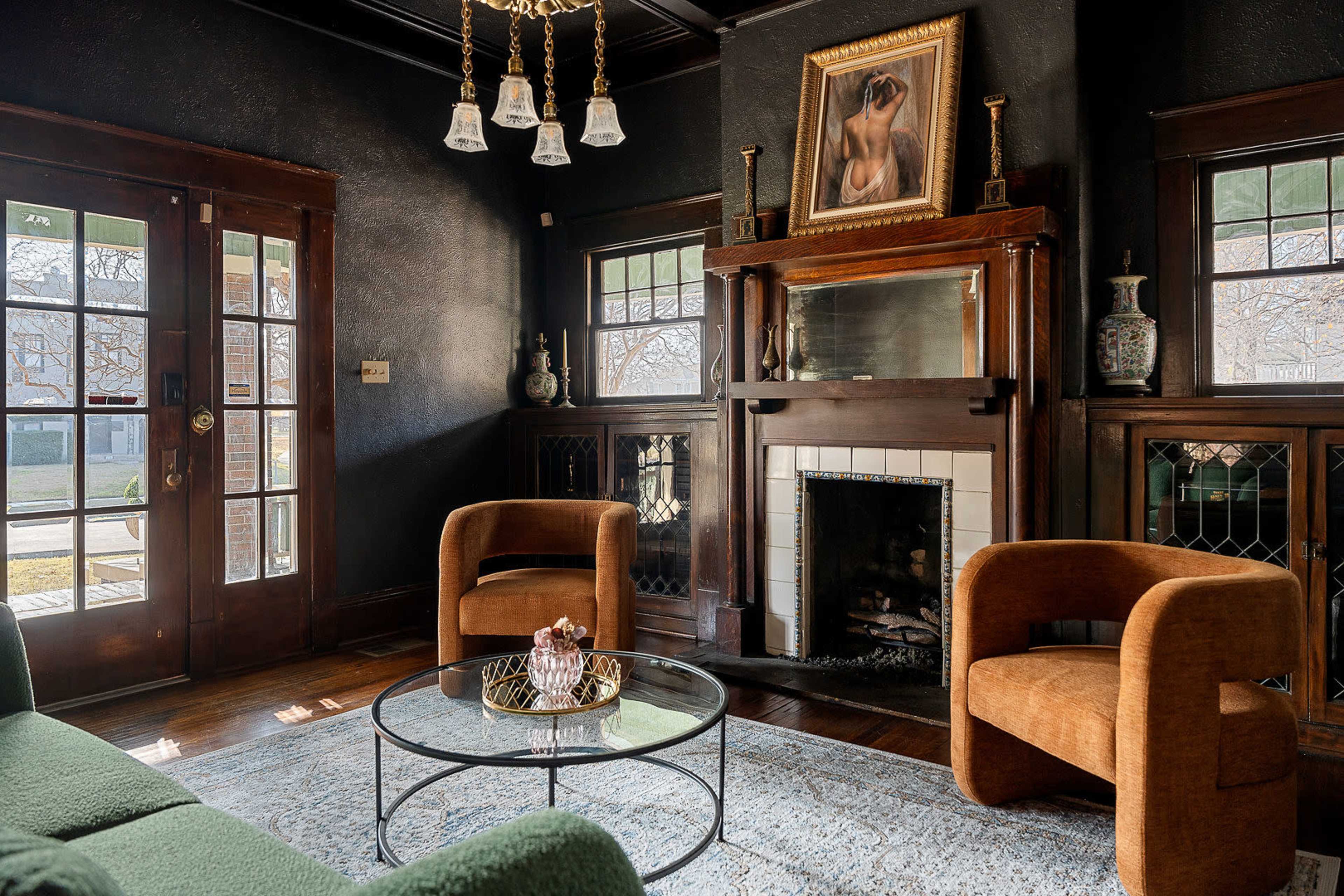The image shows a living room featuring a green sofa, two brown chairs, a glass coffee table, and a fireplace with a painting above it.