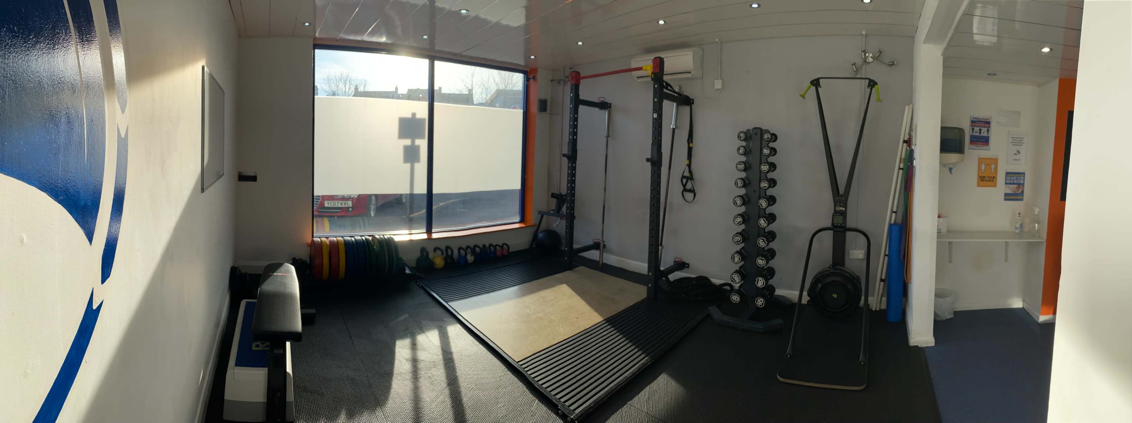 The image shows a fitness gym with various equipment including weights, a squat rack, and a mat area, all illuminated by natural light from a large window.