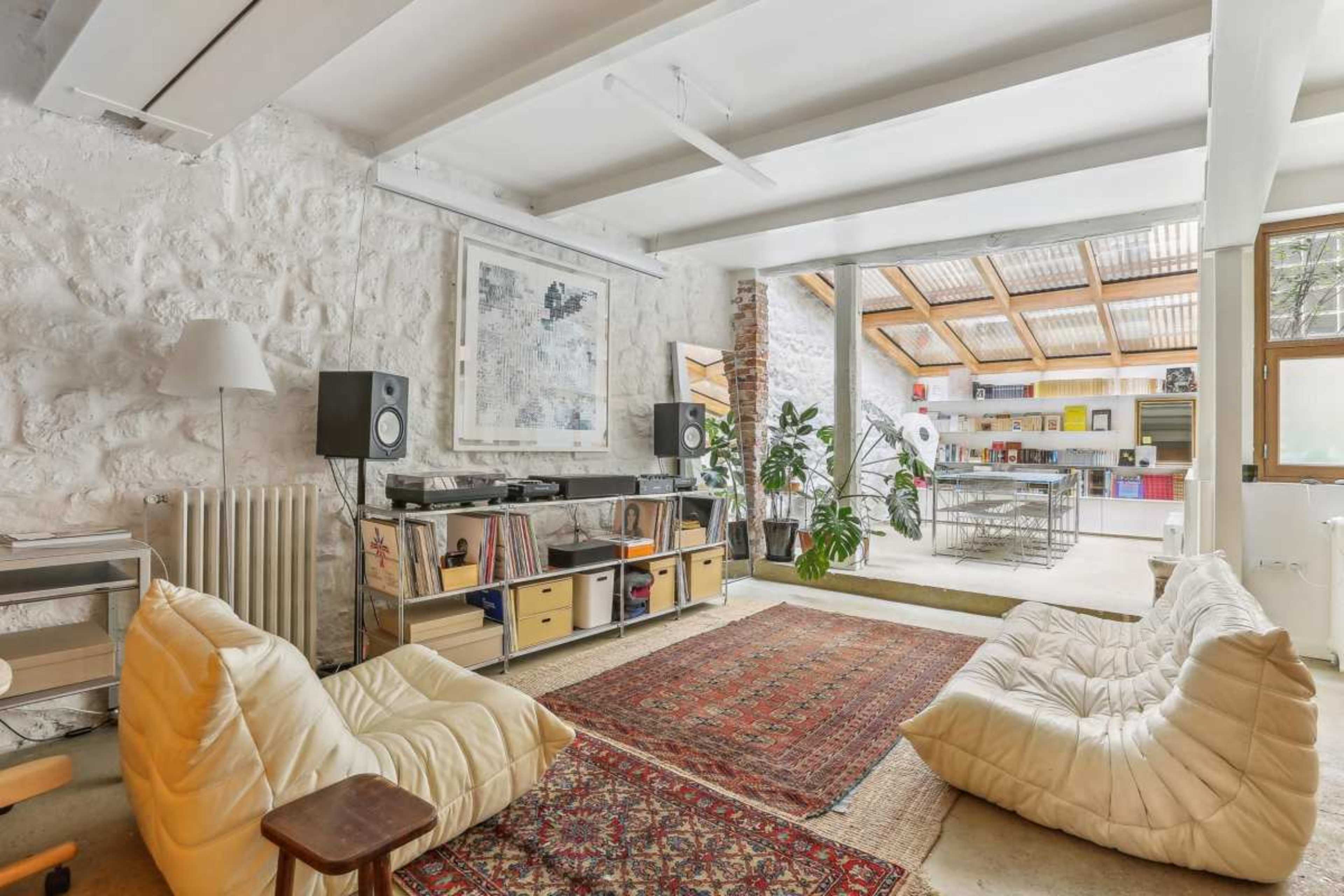 The image features a spacious, modern living area with white stone walls, low seating, a record player, and large windows that connect to a well-lit kitchen.