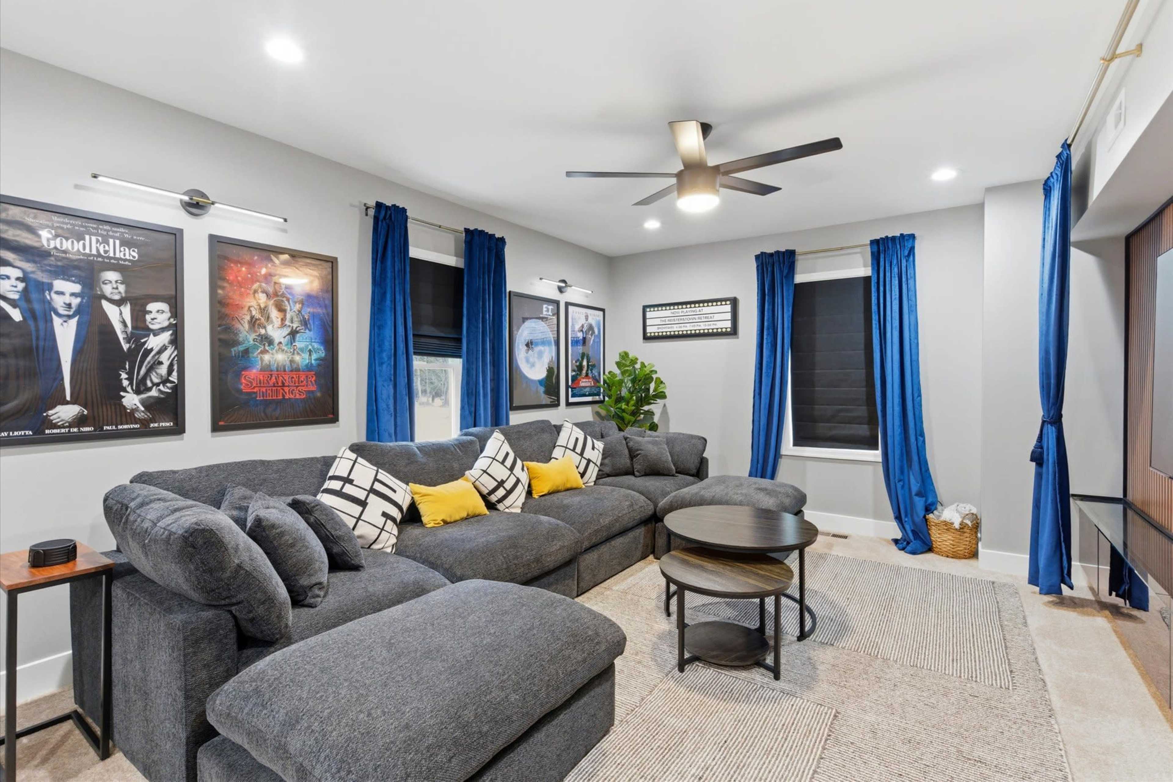 The image shows a modern living room with a gray sectional sofa, blue curtains, movie posters on the walls, and a round coffee table.