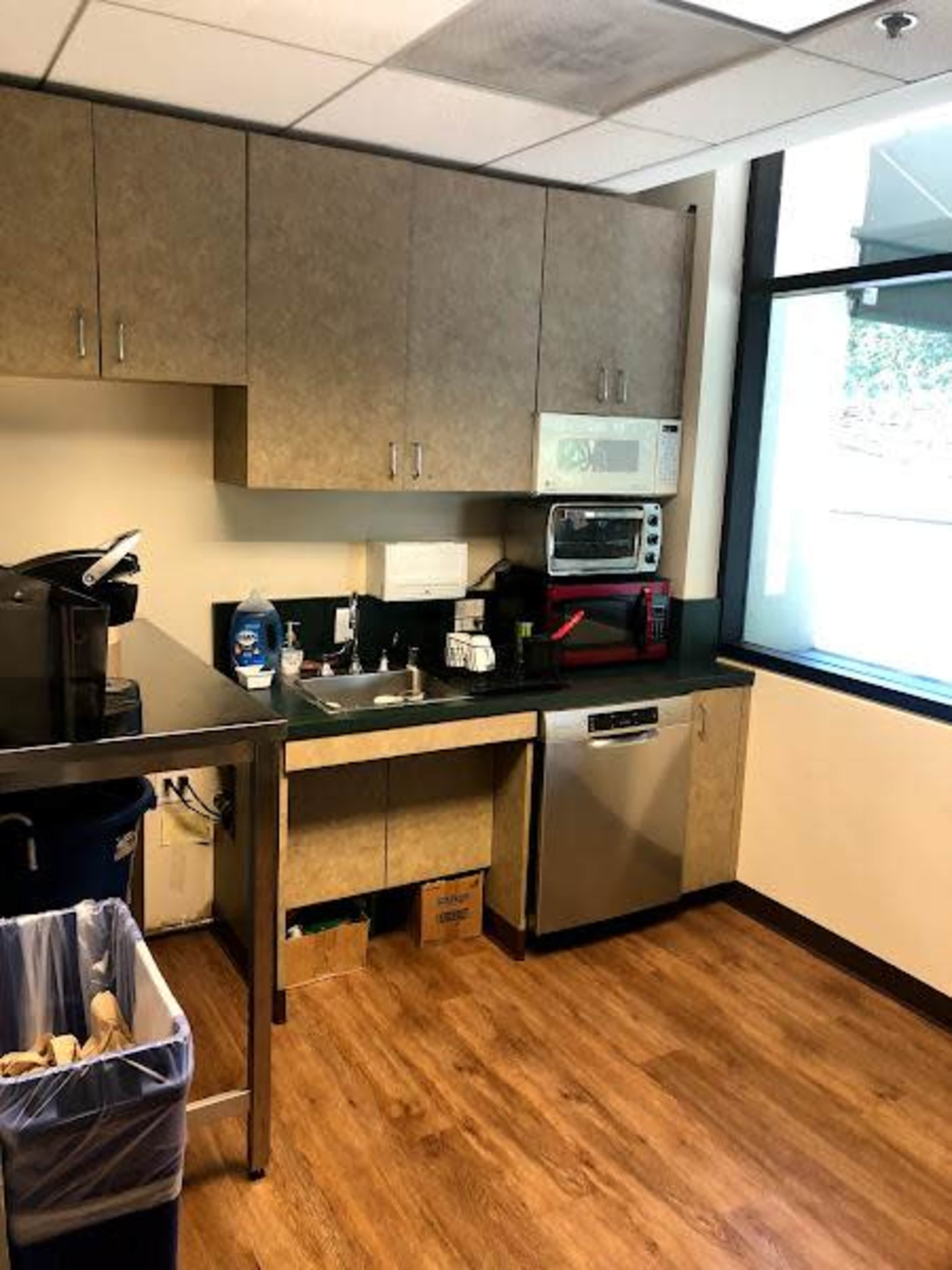 A kitchenette with cabinets, a sink, a microwave, a coffee maker, and a dishwasher, beside a trash bin.