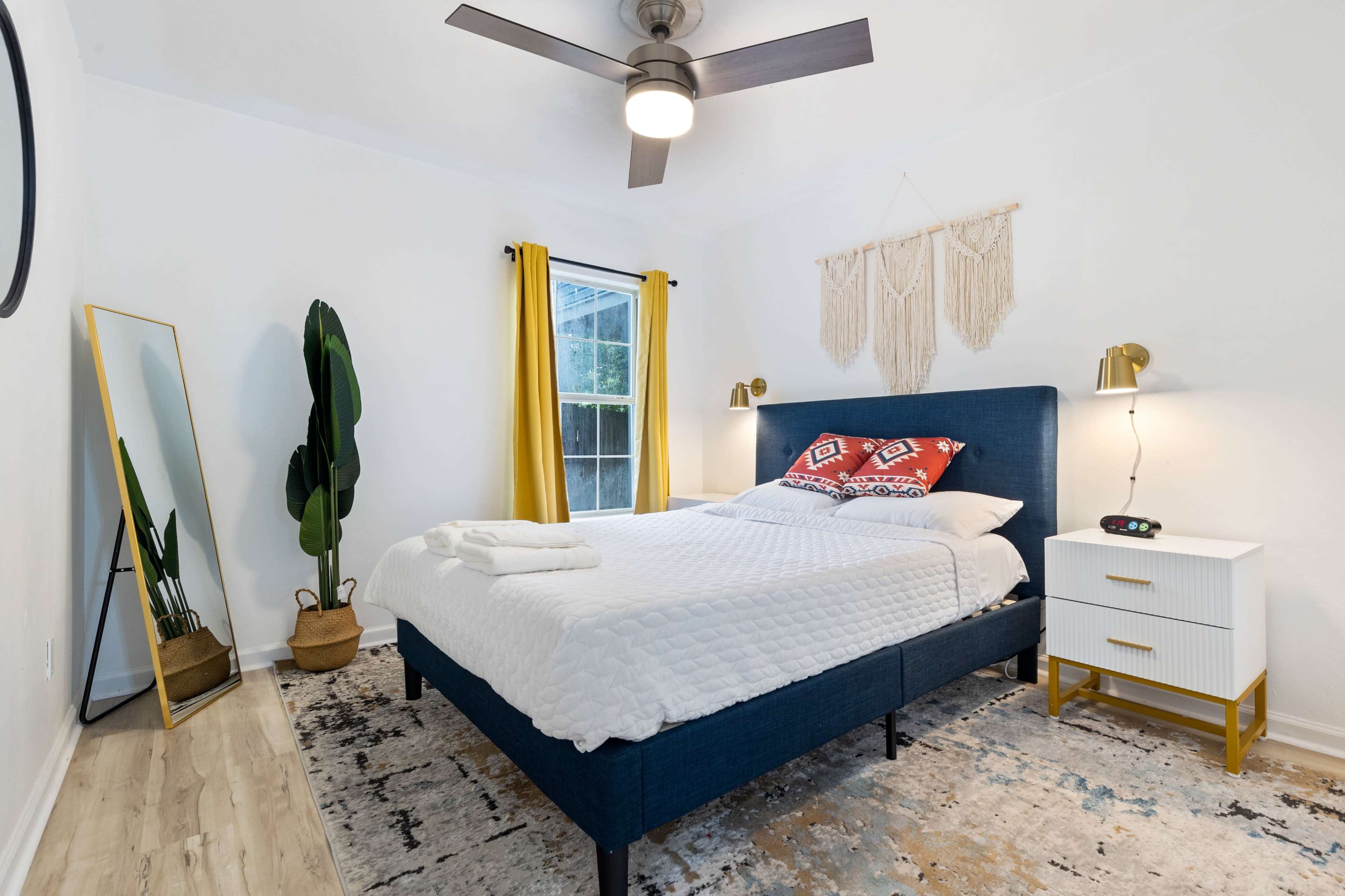 A neatly arranged bedroom features a blue bed with decorative pillows, a tall plant in a pot, yellow curtains, and a large mirror against the wall.