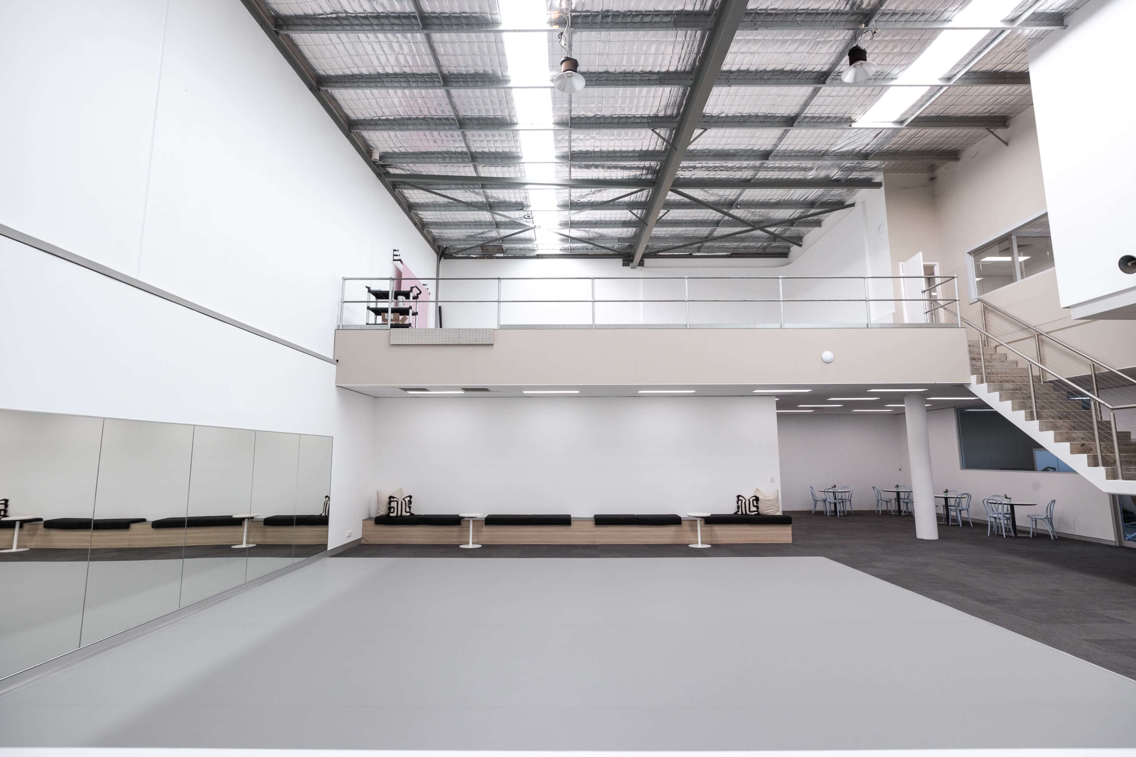 A spacious, brightly lit dance studio with a mirrored wall, a gray floor, and a mezzanine level overlooking the main area.