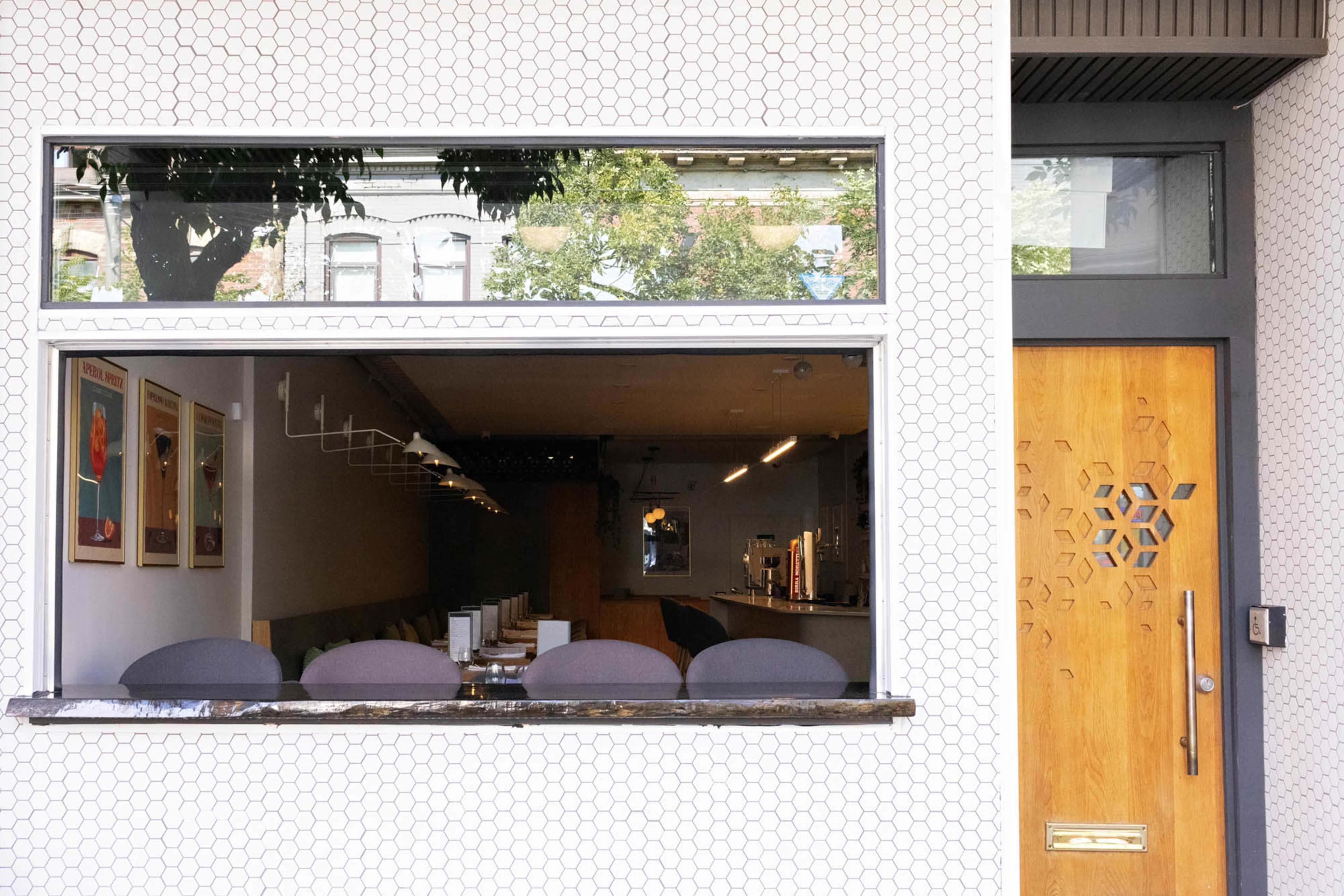 The image shows a modern restaurant exterior with a hexagonal-patterned wall, featuring a large window that reveals a minimalist interior and a wooden door with a geometric design.