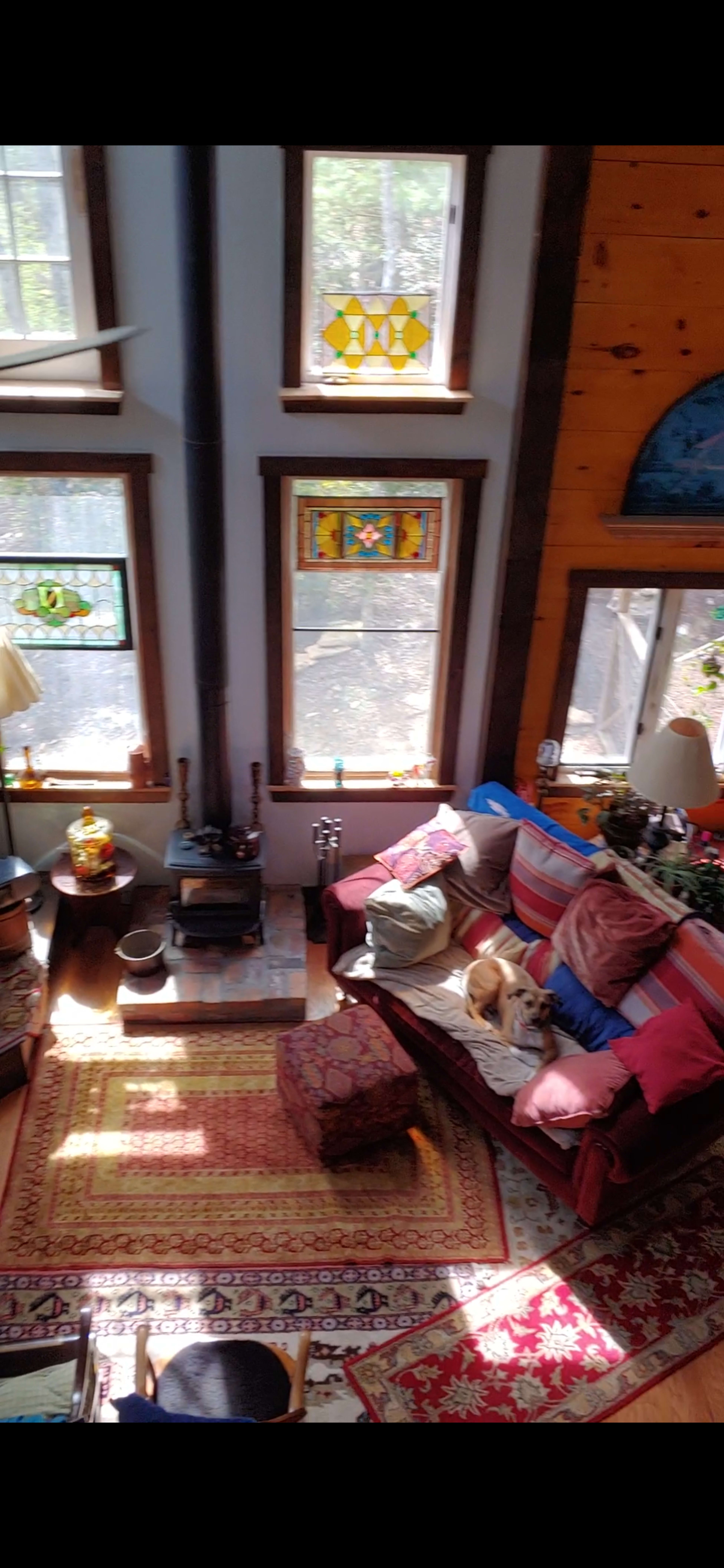 The image shows a cozy living room with colorful stained glass windows, a wood stove, and a sofa accompanied by a dog resting on it.
