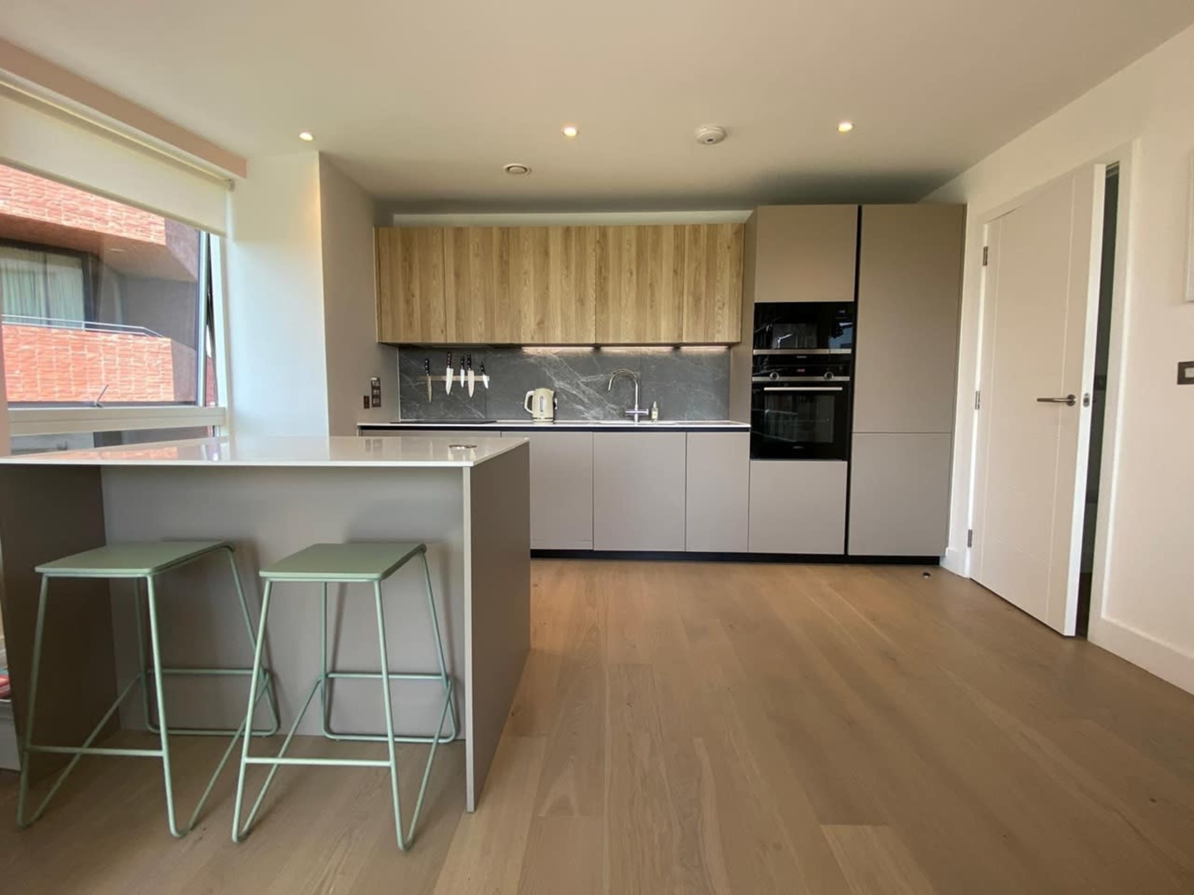The image shows a modern, minimalist kitchen featuring wooden cabinetry, sleek gray countertops, and two green stools by an island.