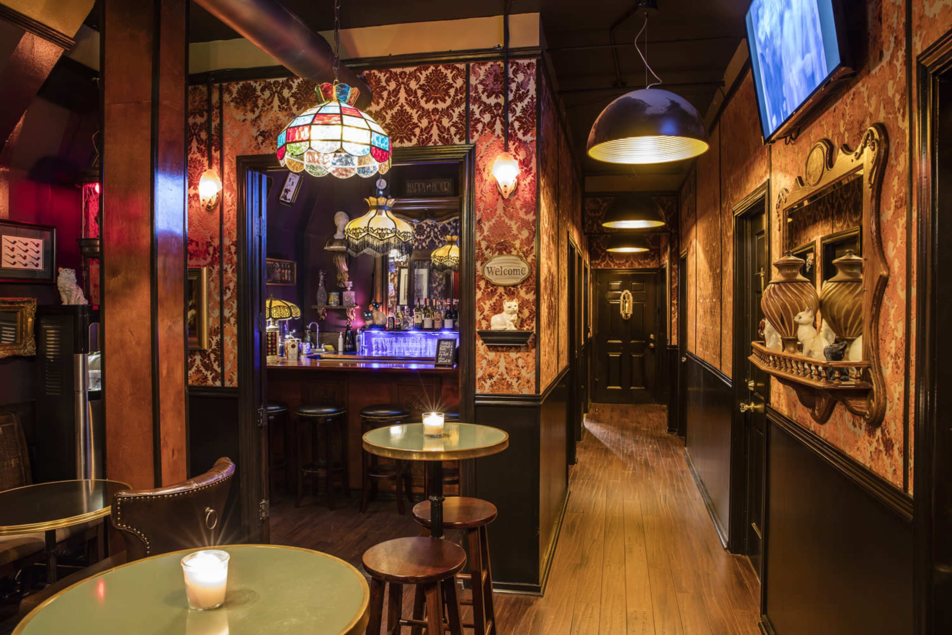 A dimly lit bar corridor with decorative wallpaper, a stained glass lamp, and bar stools lining the walls.