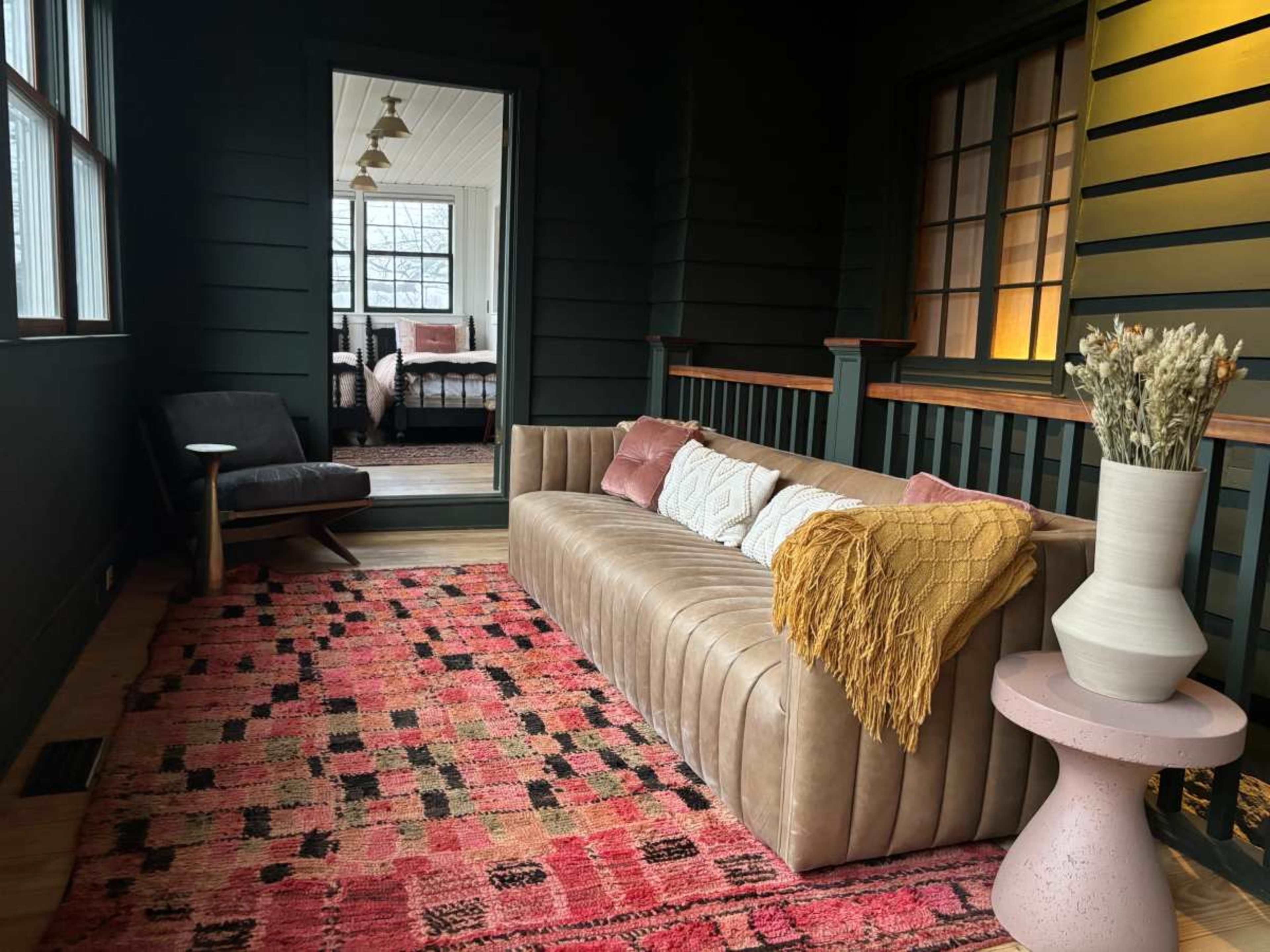 The image shows a furnished porch with a leather sofa, decorative pillows, a pink side table, and a patterned rug, leading to a brightly lit room in the background.