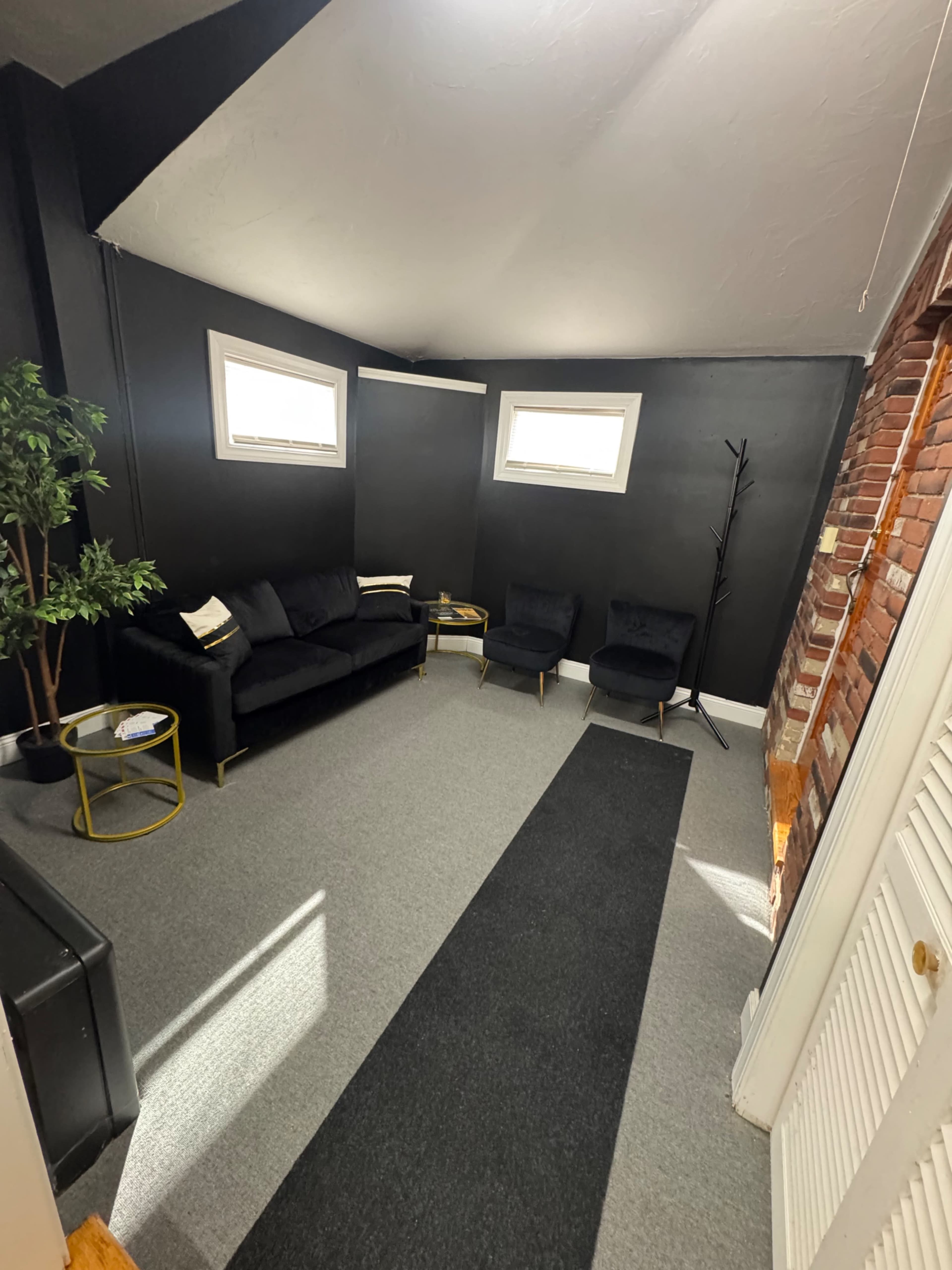 The image shows a small room with dark gray walls, two black armchairs, a black sofa, a small side table, and a potted plant in one corner.