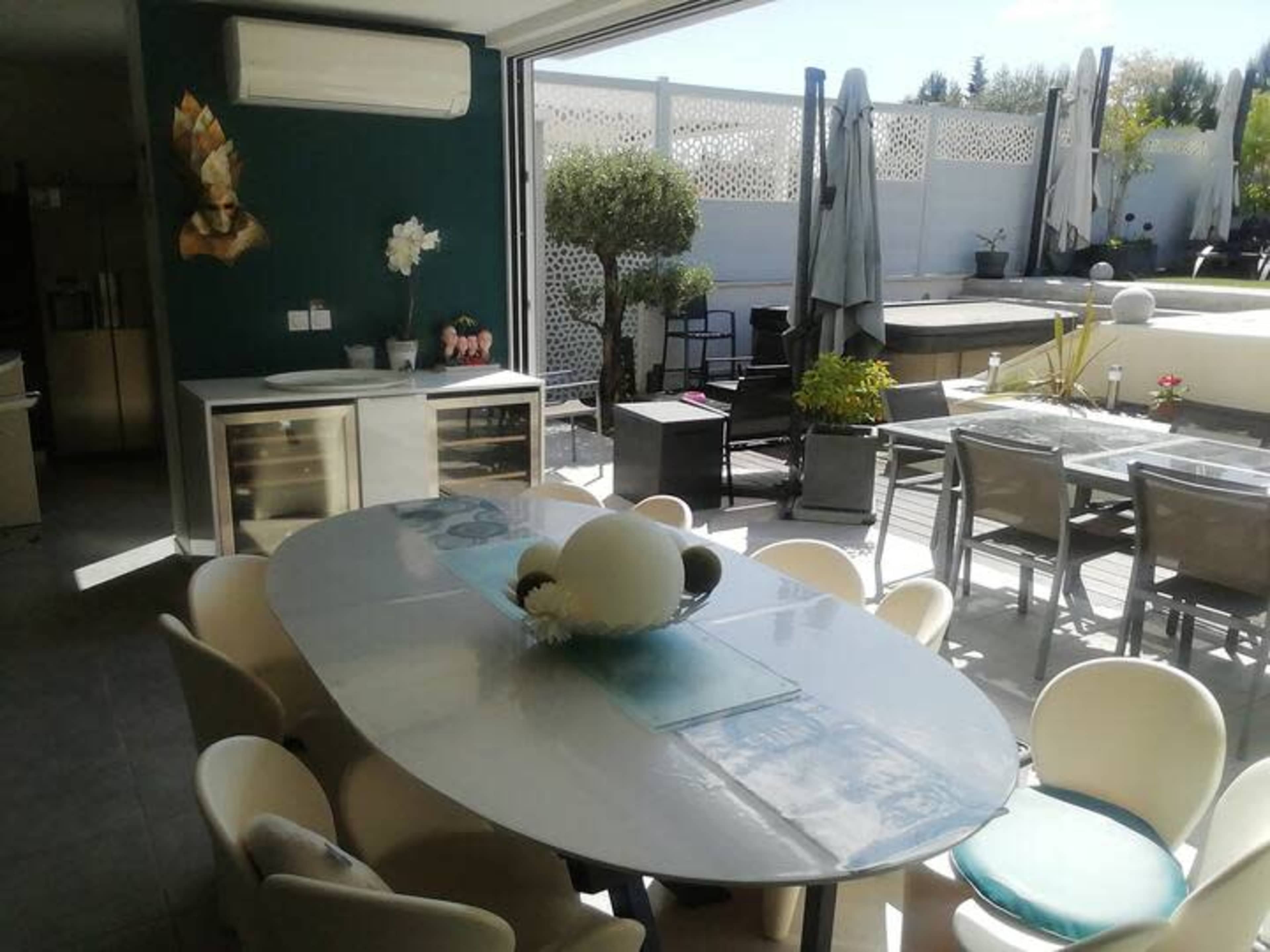 The image shows a modern dining area with a large oval table surrounded by chairs, overlooking an outdoor seating space with plants and a sun umbrella.