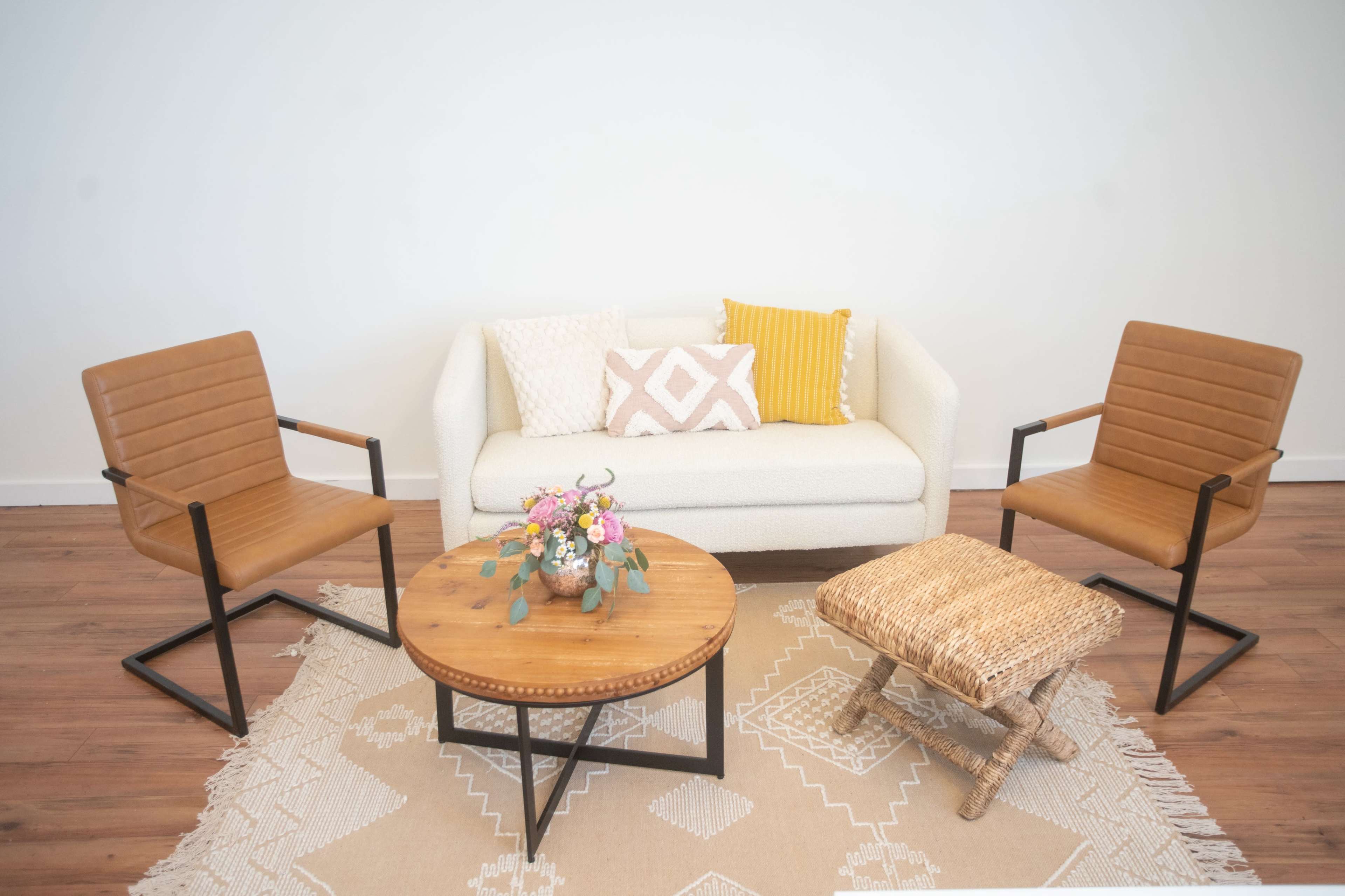 The image features a cozy living room setup with a white sofa, two brown chairs, a wooden coffee table, a woven ottoman, and a floral centerpiece on the table, all arranged on a decorative rug.