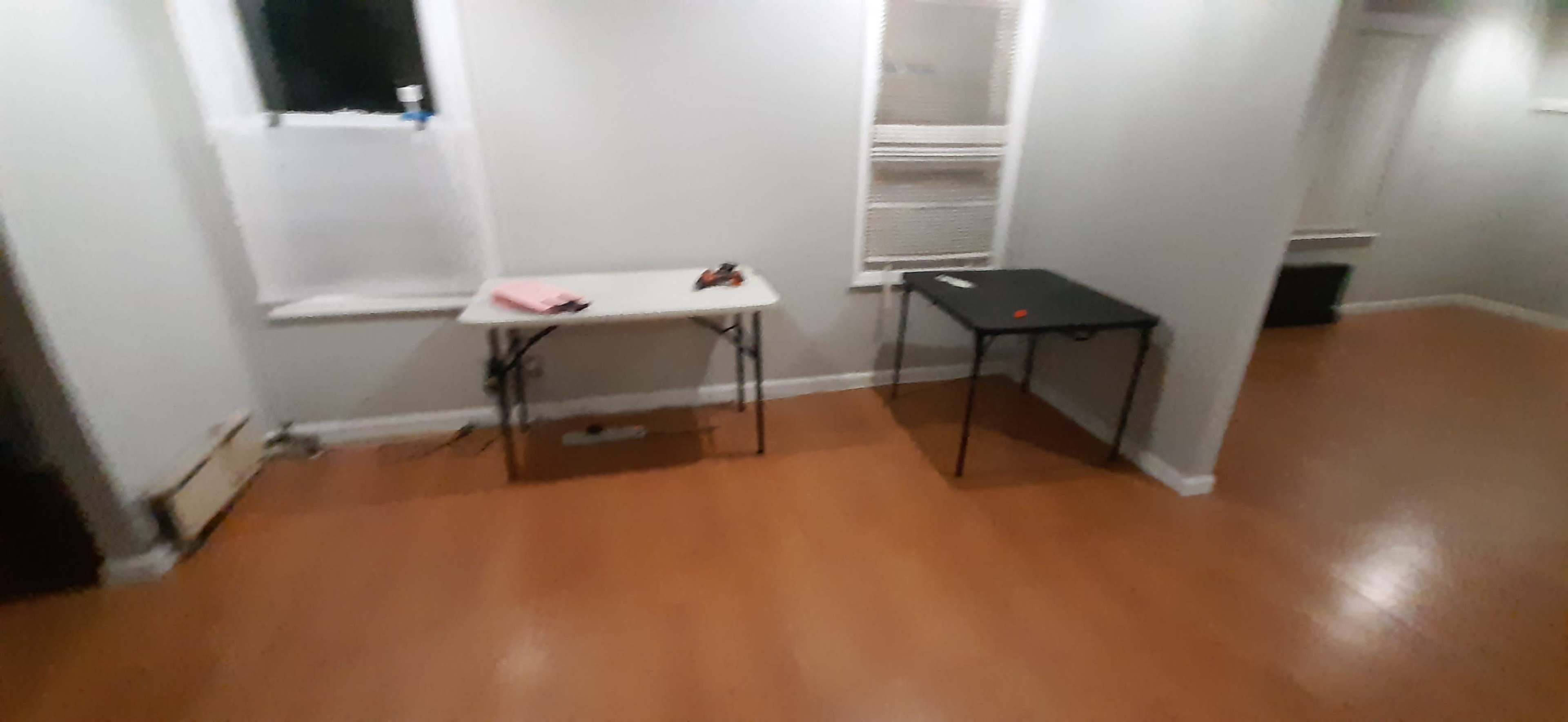 A small, empty room with light-colored walls and wooden flooring, featuring two folding tables on opposite sides.