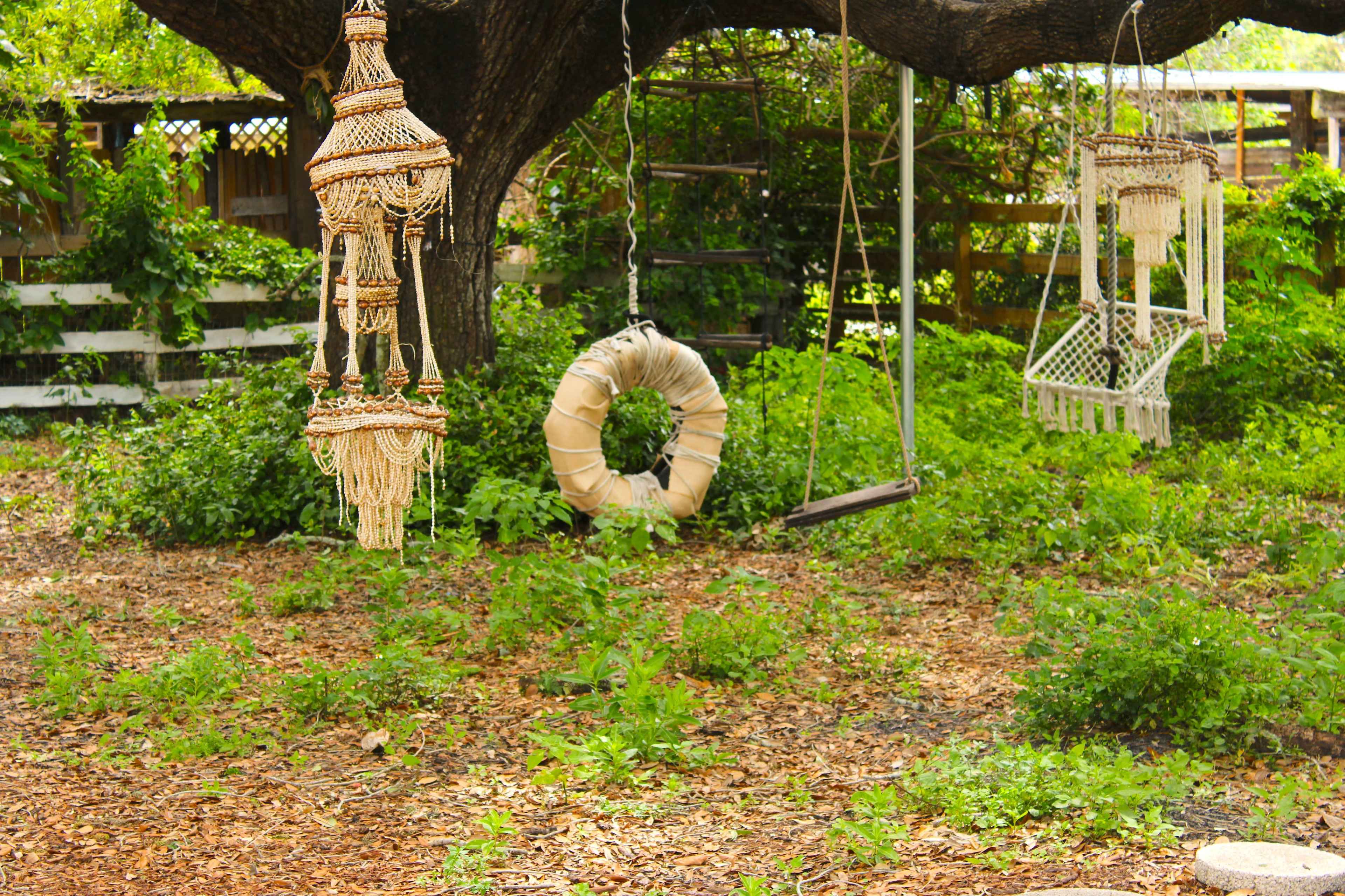 The scene features a tree with macramé hangings, a swing, and a round decoration resembling a life ring, set in a garden with lush greenery and mulch underfoot.