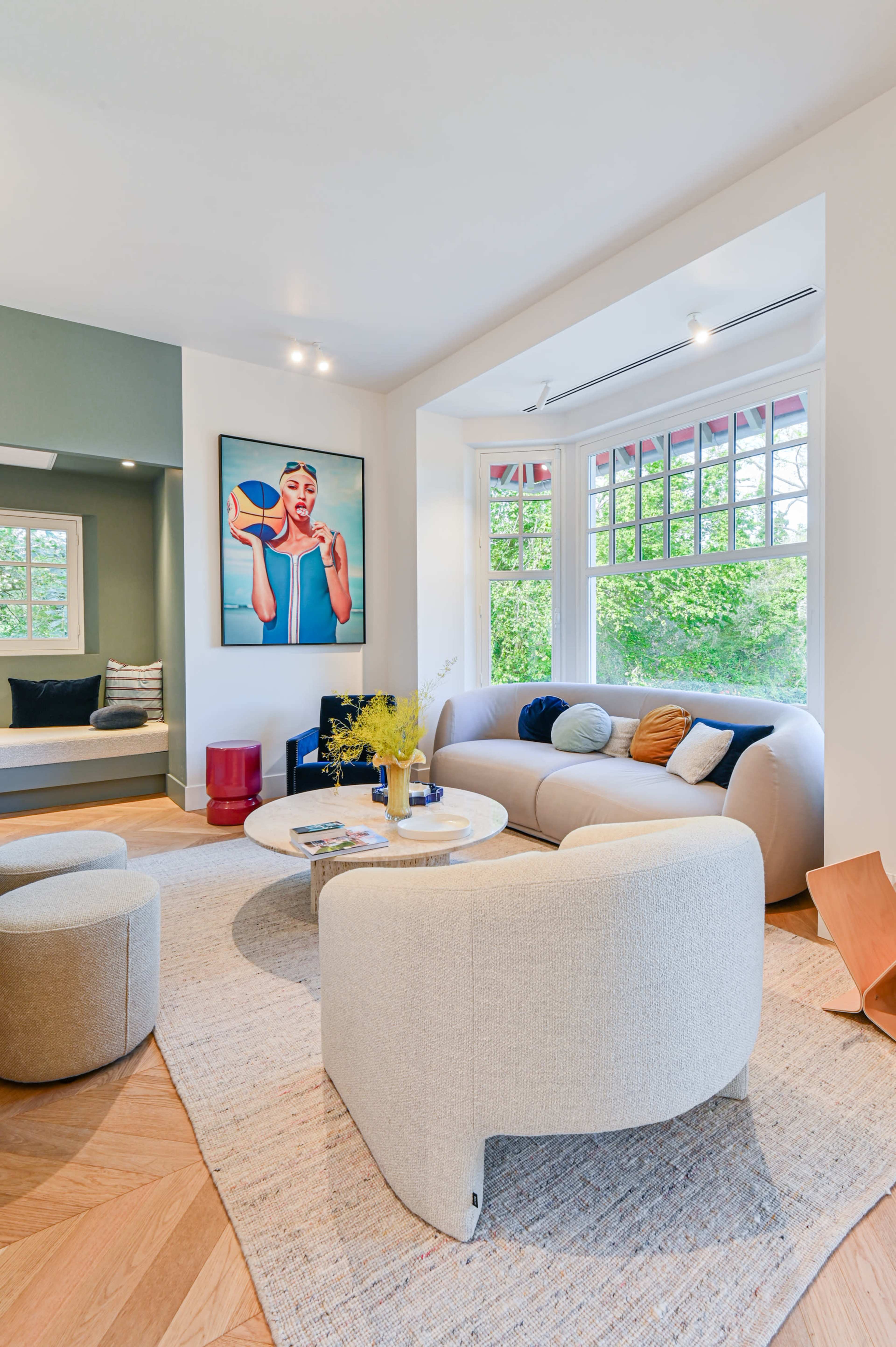The image shows a modern living room featuring a curved sofa, circular chairs, a coffee table, and a large artwork on the wall, with greenery visible through the windows.