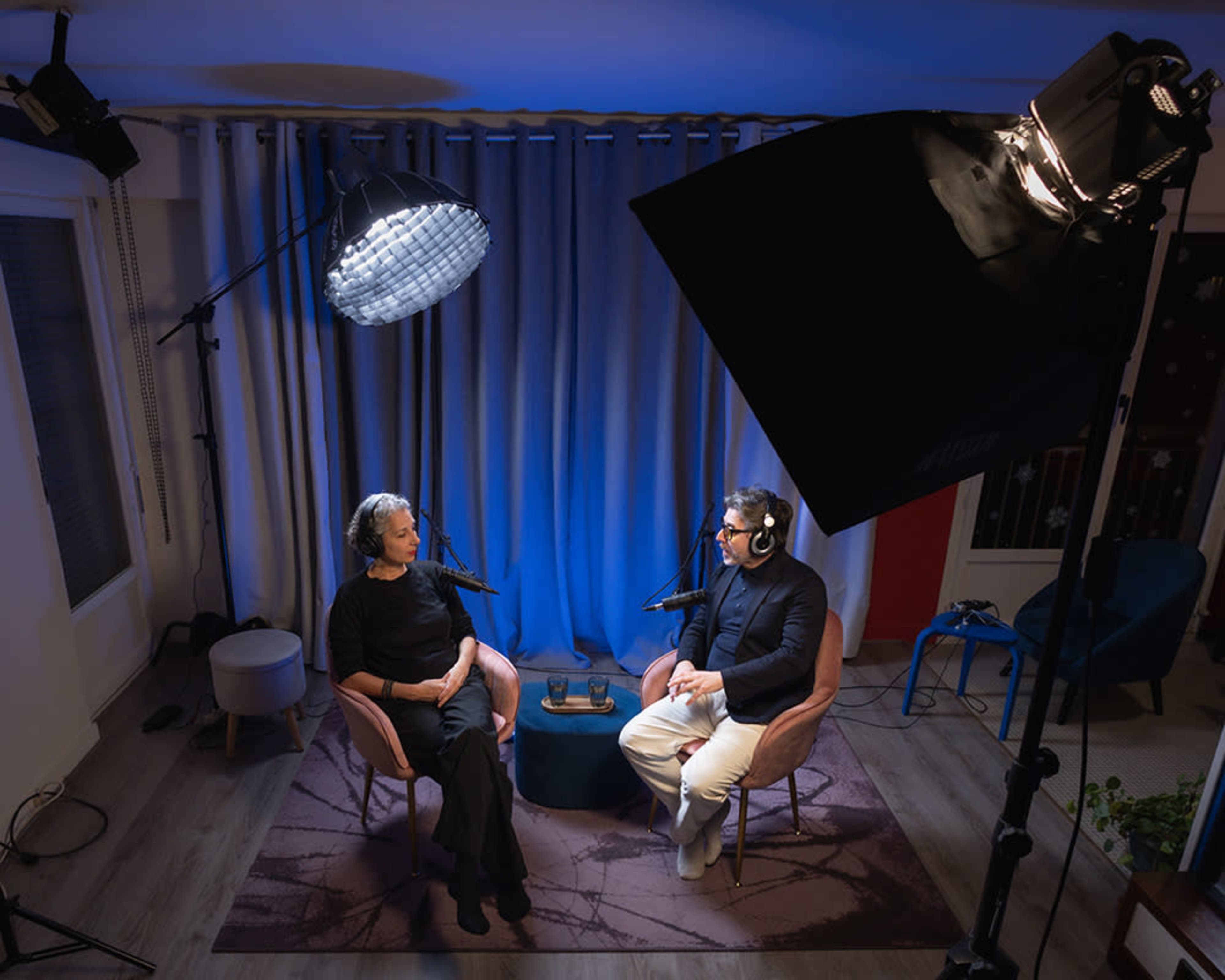 A woman and a man sit across from each other in a podcast studio, each wearing headphones and speaking into microphones, with blue lighting illuminating the scene.