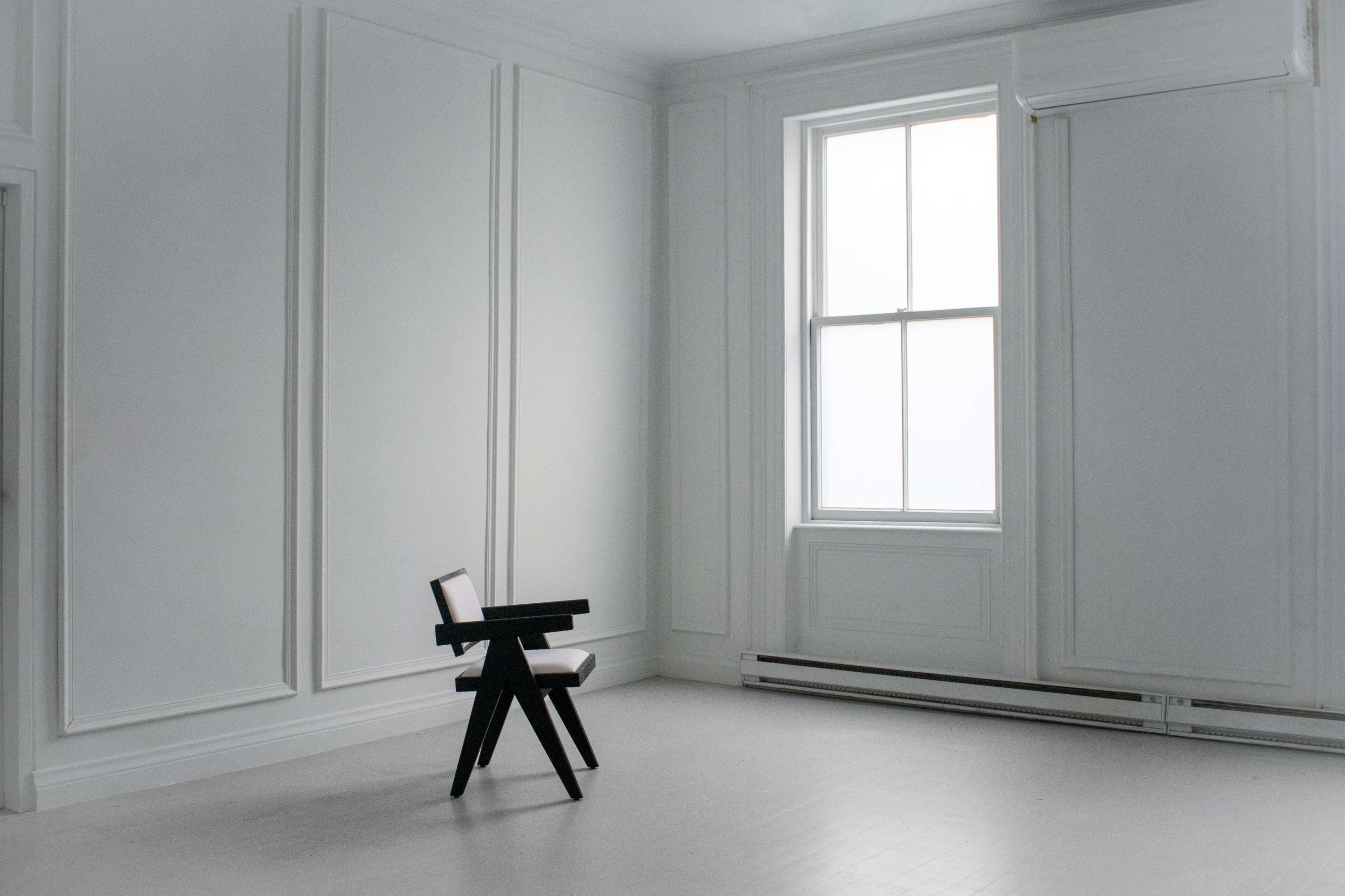 A minimalist room with white walls, a large window, and a single black chair located in the center.
