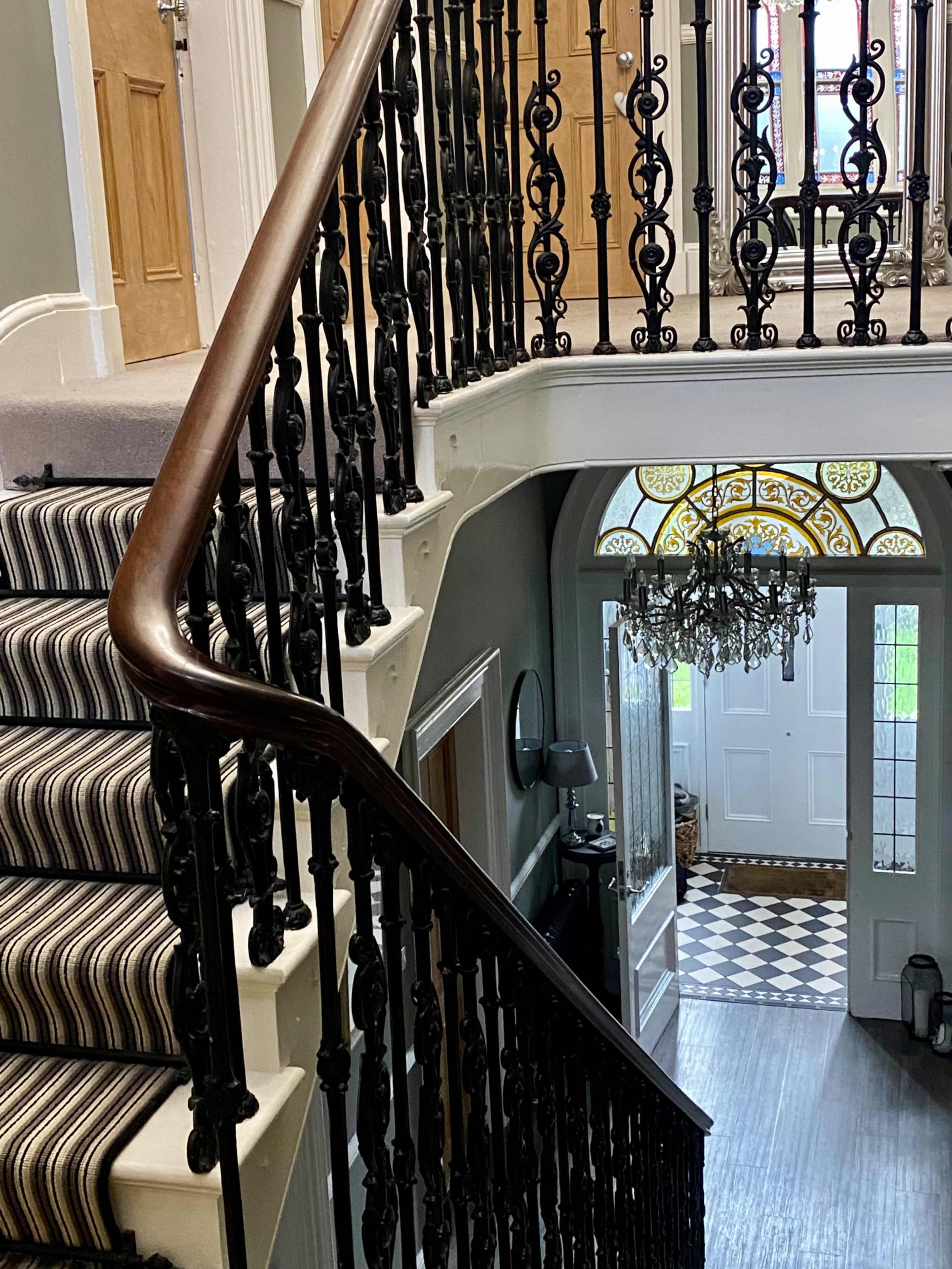 A staircase with a wooden handrail and decorative wrought iron balustrade, leading to an entrance hallway featuring a patterned floor and a chandelier.