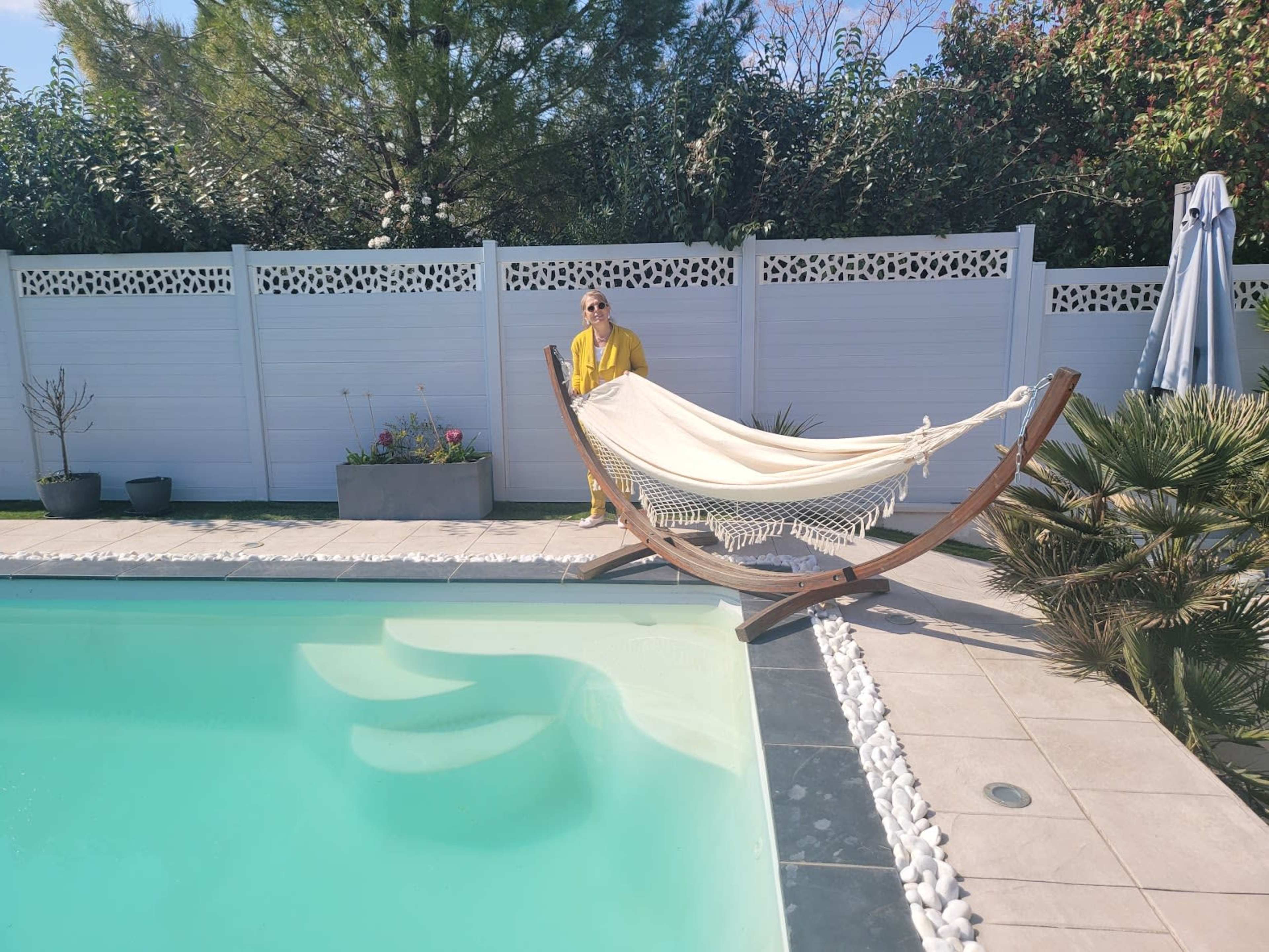 A person in a yellow outfit stands near a hammock beside a swimming pool, with a landscaped area and a white fence in the background.