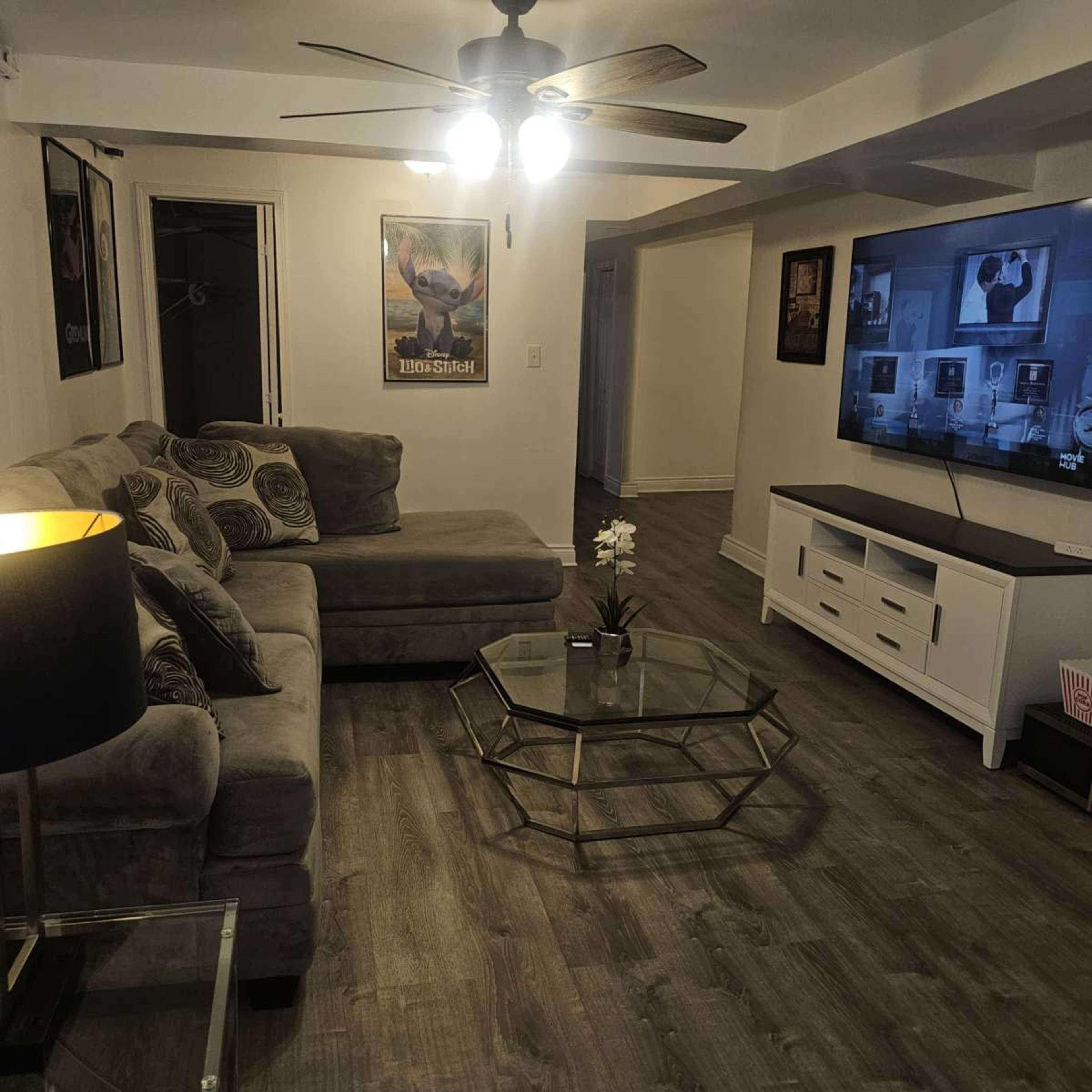 The image shows a living room with a sectional sofa, a coffee table, a ceiling fan, and a television mounted on the wall.