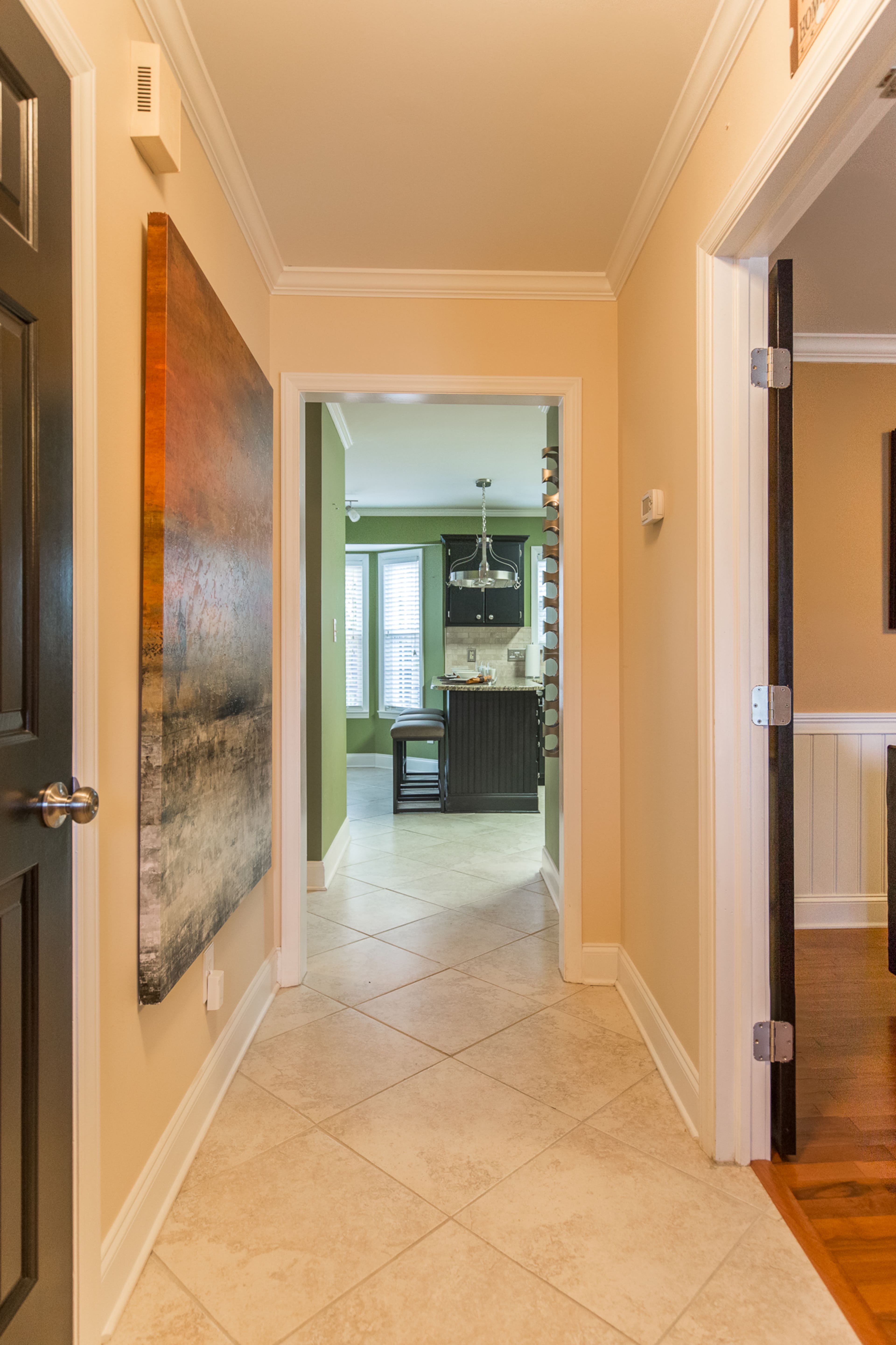 A hallway with beige walls and tile flooring leads to a kitchen visible at the end, featuring dark cabinetry and natural light through windows.