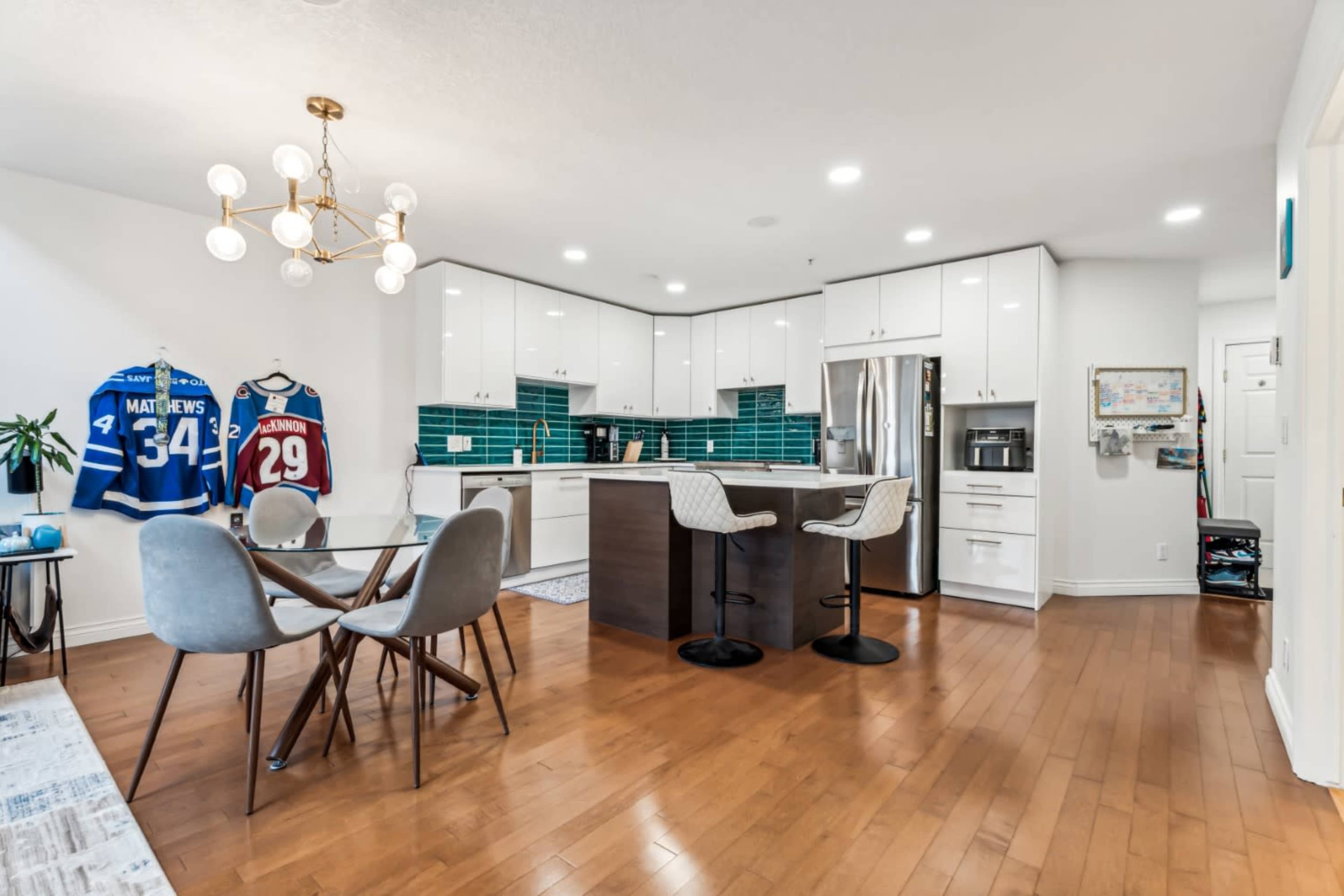 The image shows a modern kitchen and dining area featuring a round glass table, four chairs, and sports jerseys displayed on the wall.