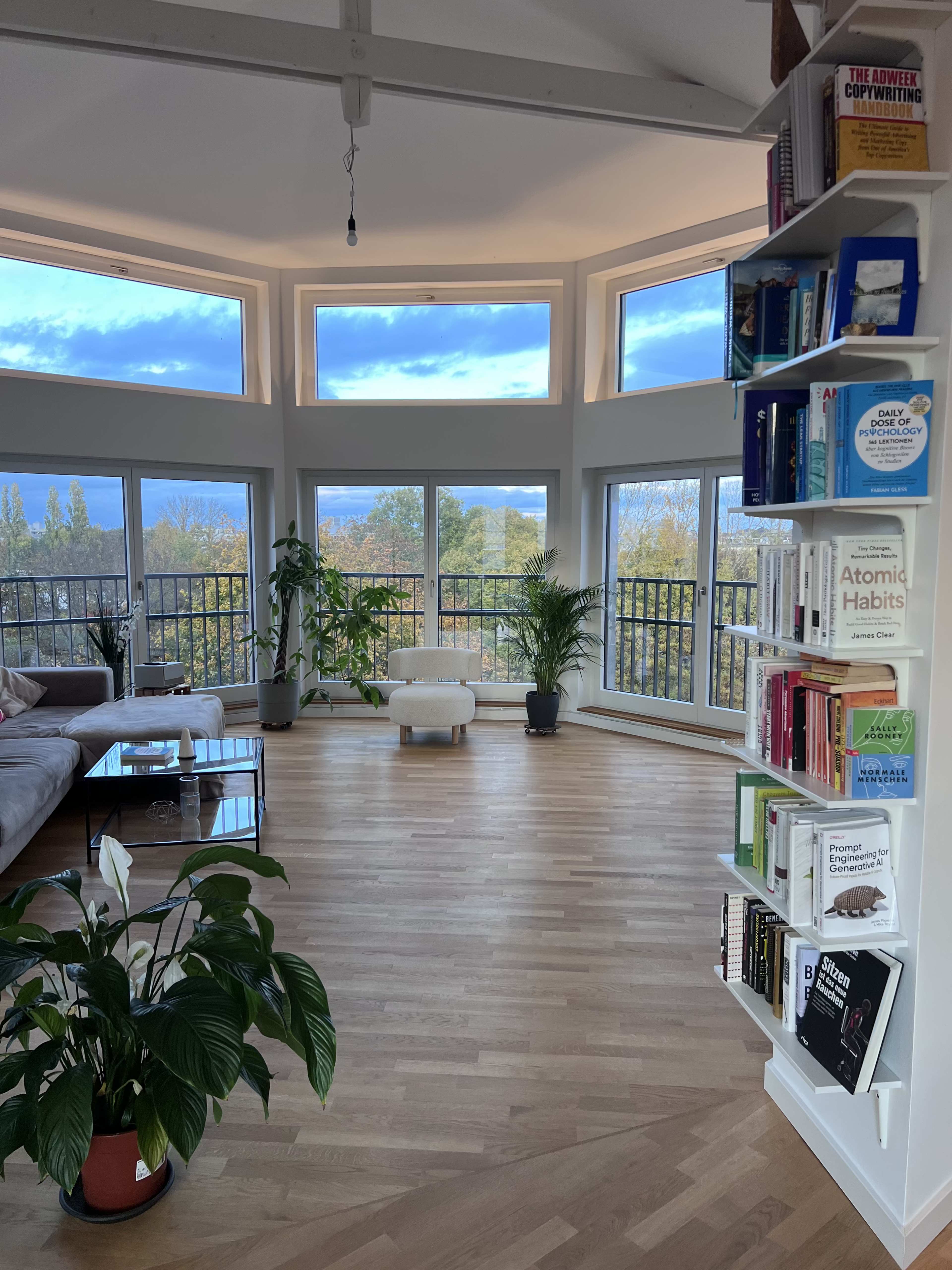 A bright and modern living room features large windows with a view, a white lounge chair, a bookshelf with colorful books, and a potted plant.