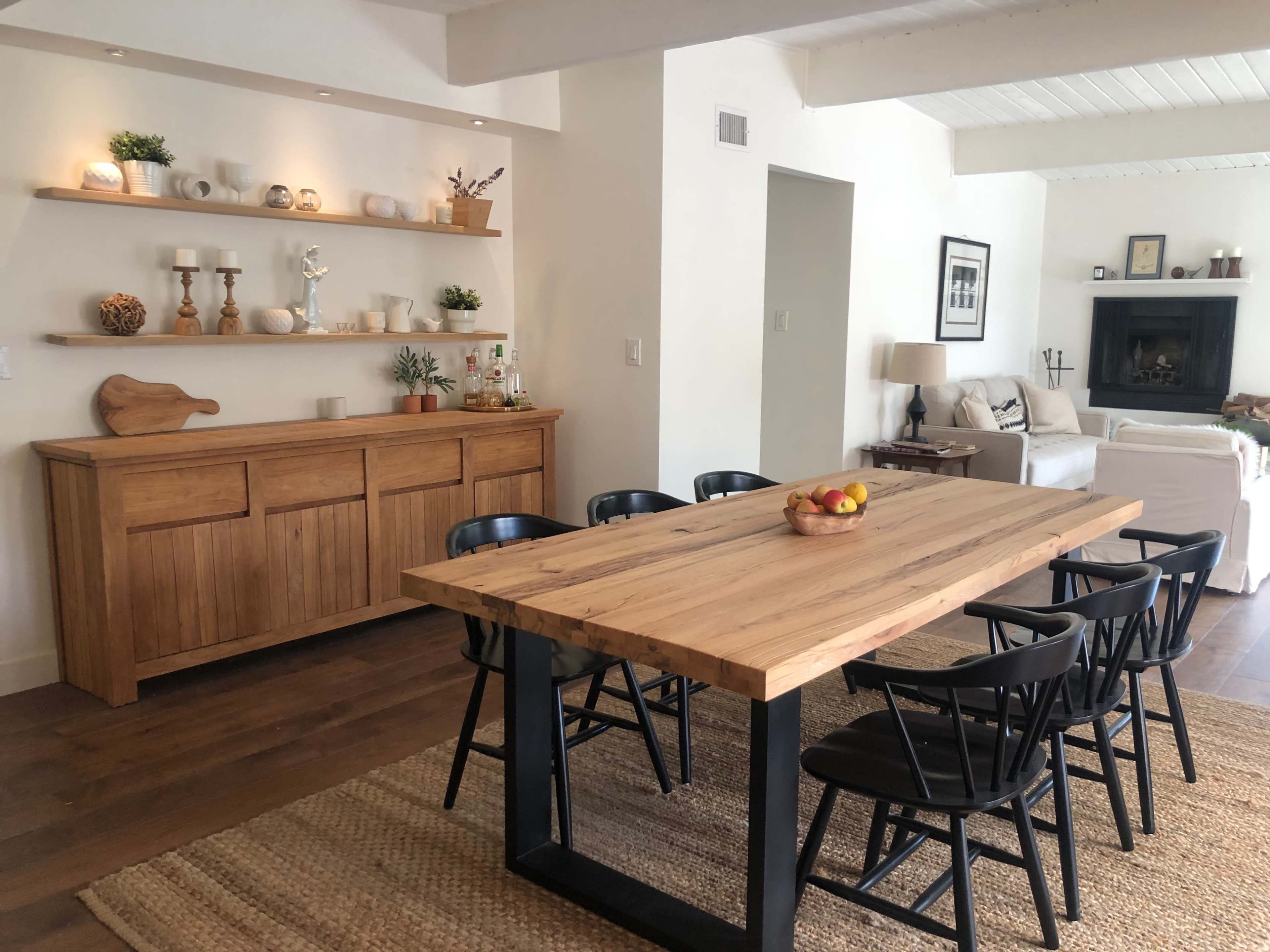 A wooden dining table with black chairs is positioned in a room featuring a sideboard and shelves displaying decorative items.