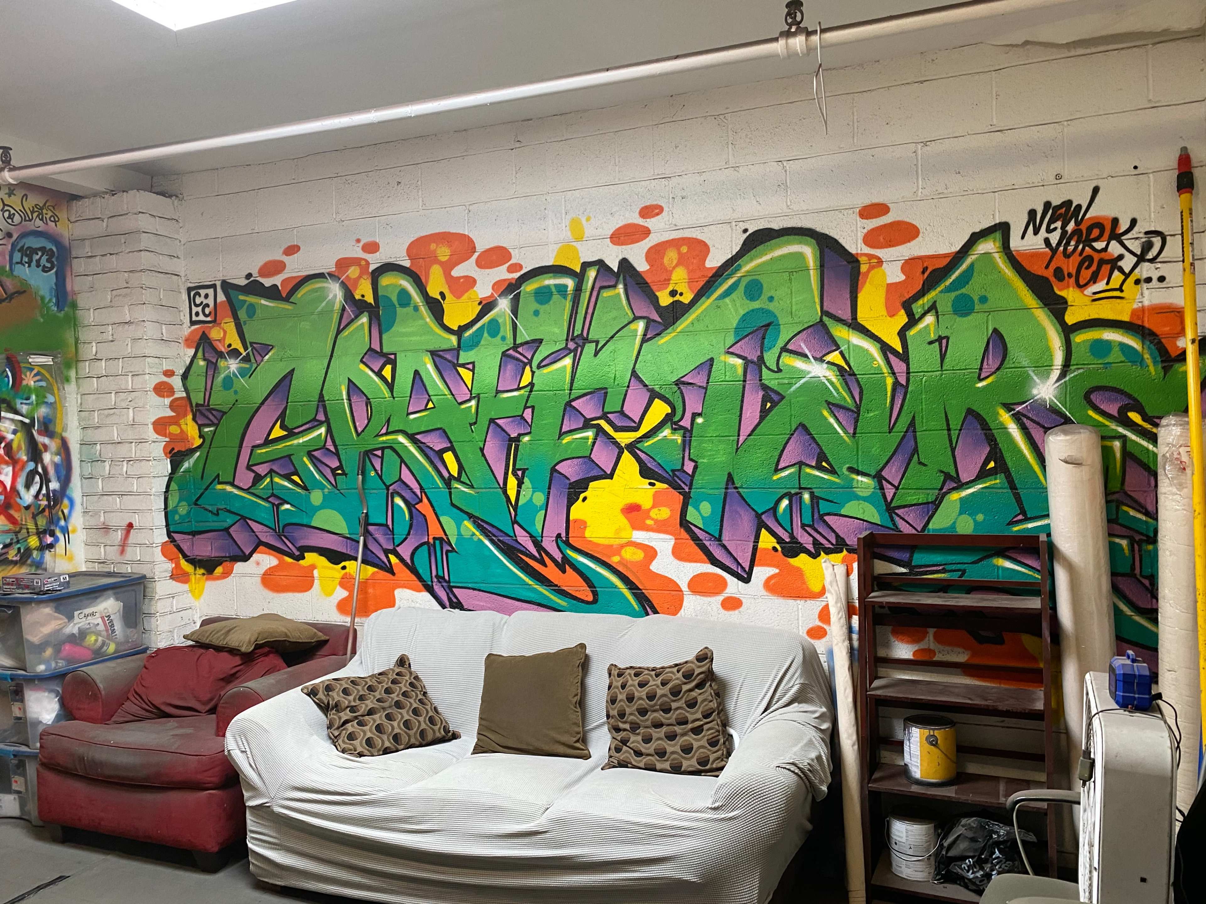 A vibrant graffiti mural on a wall, with the word "GRAFFITI" displayed in bold, colorful letters, alongside a red and orange splatter background, with a white sofa in front.