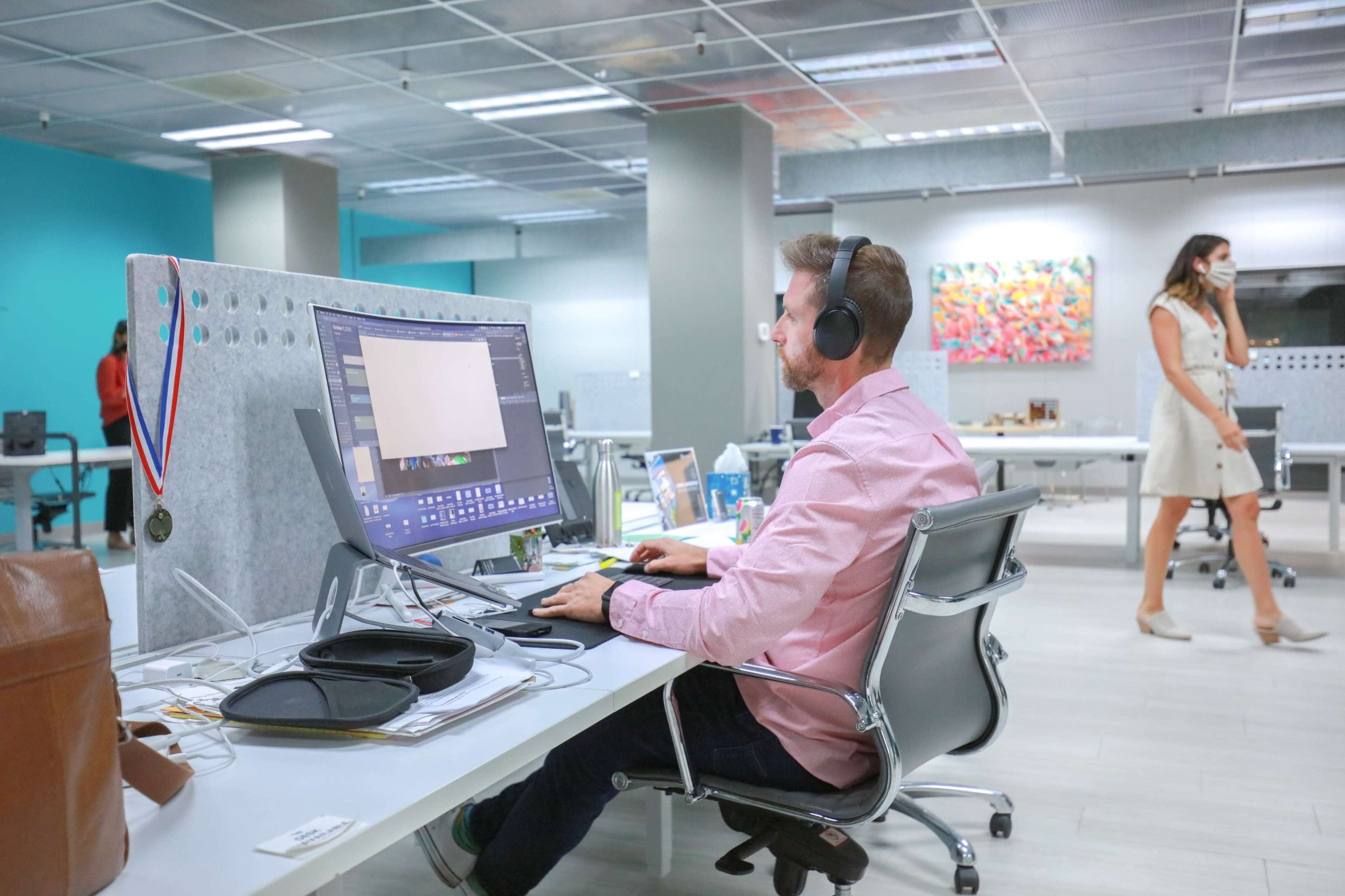 A man wearing headphones works at a desk with a computer in a modern office, while a woman walks by in the background.