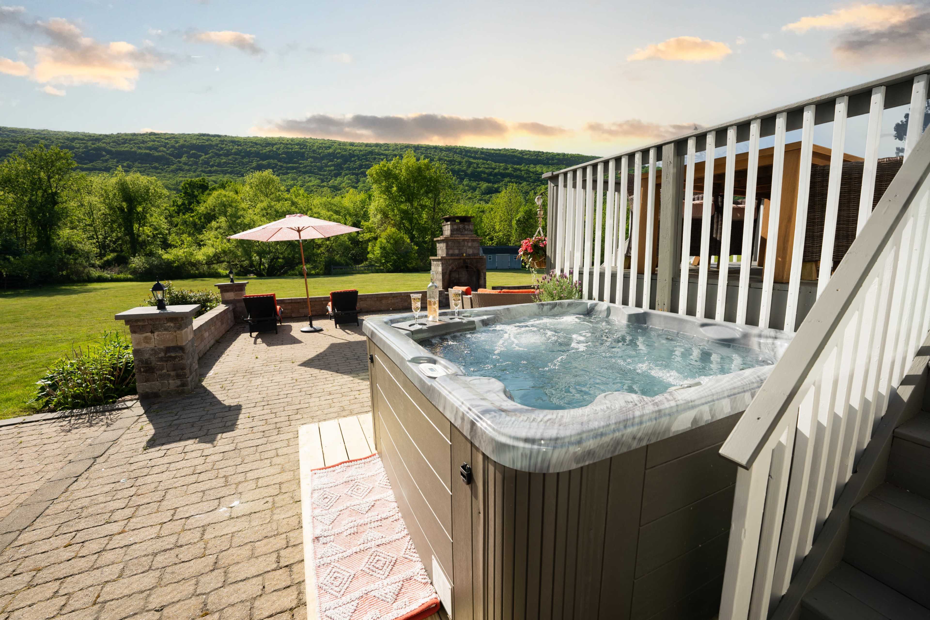 A hot tub is situated on a deck, overlooking a green landscape with rolling hills and a patio set nearby.