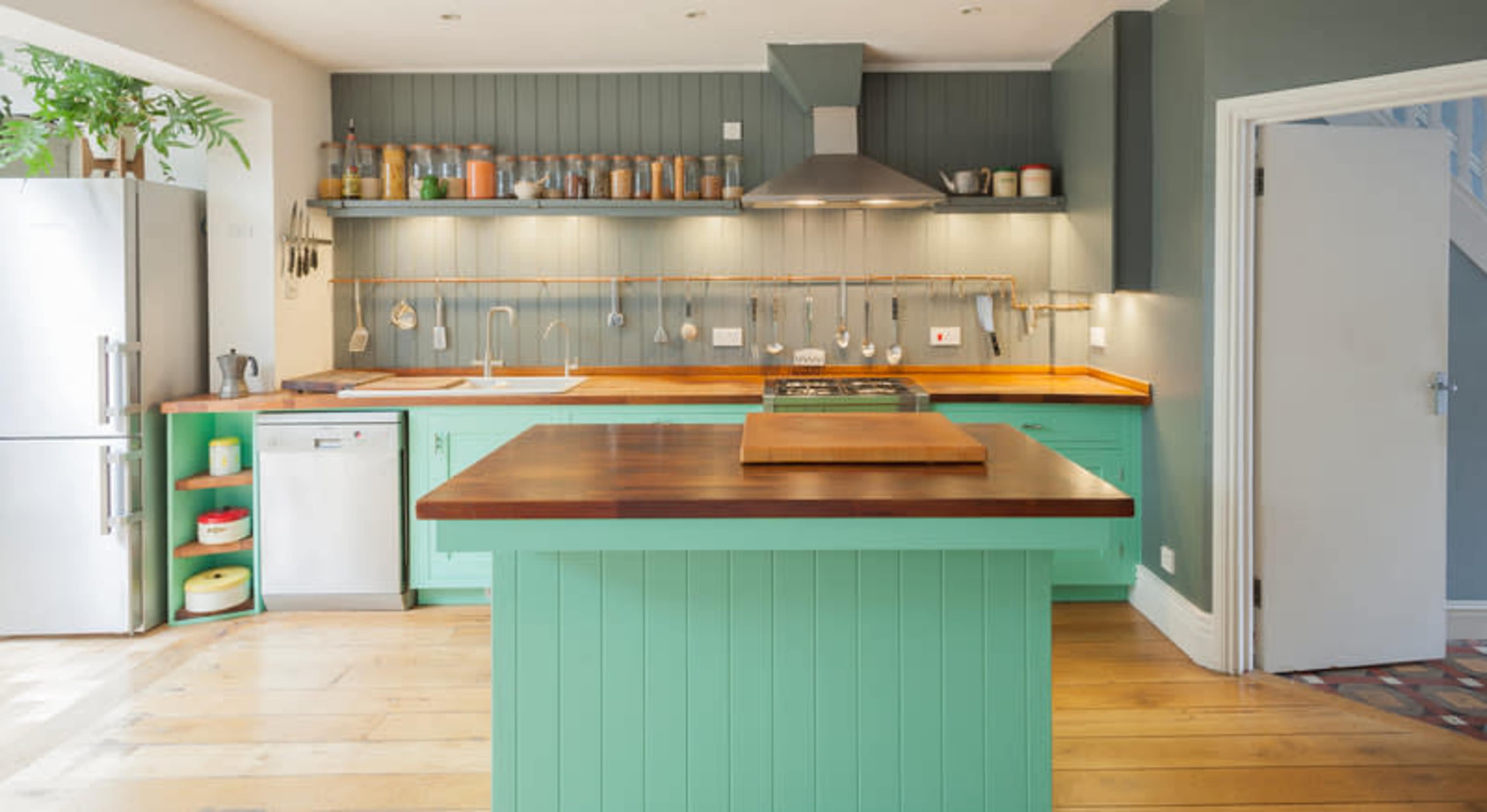 A modern kitchen with teal cabinetry, wooden countertops, and stainless steel appliances, featuring an island in the center.