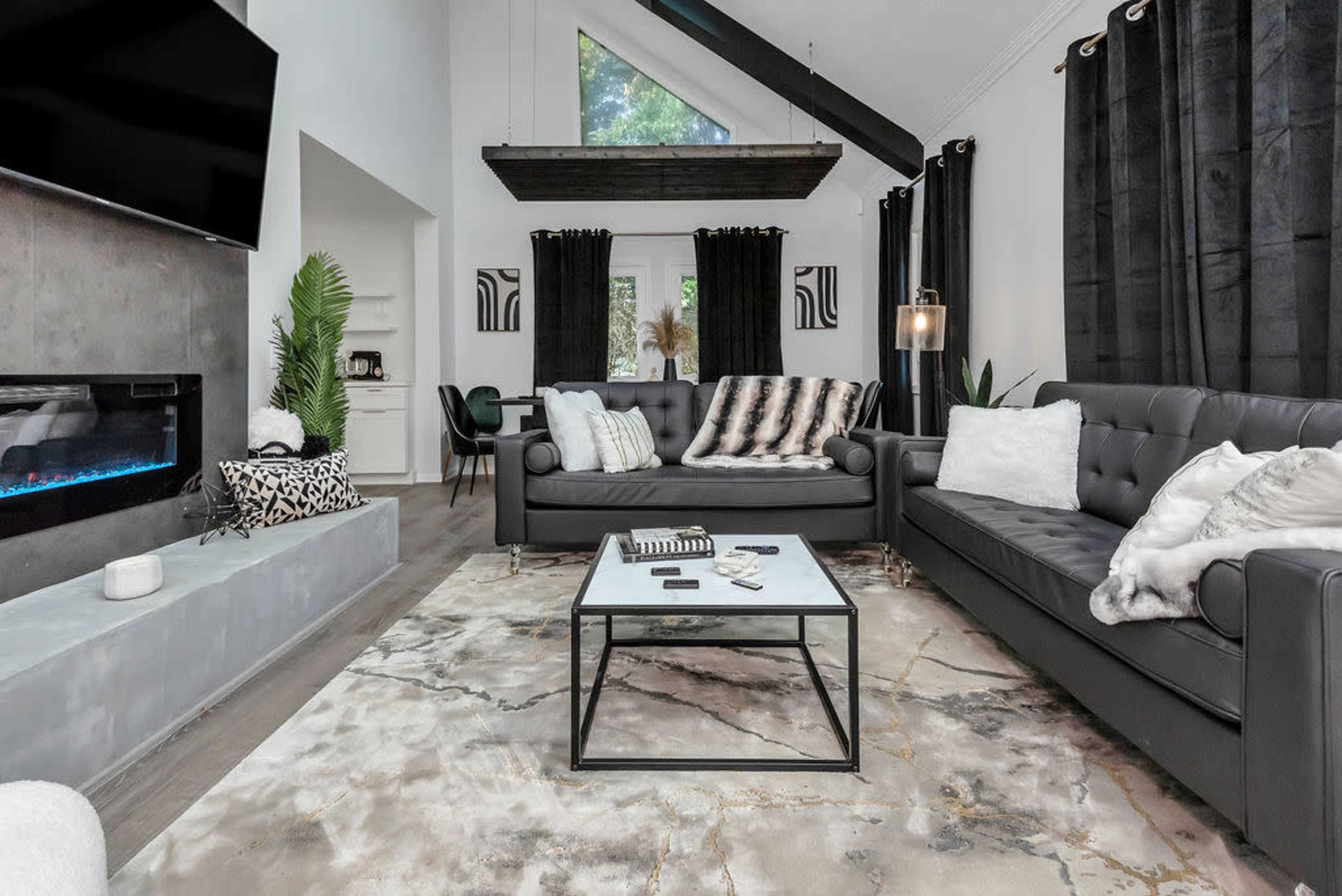A modern living room featuring a black leather sofa, a coffee table on a marble-patterned rug, and large windows allowing natural light to enter the space.