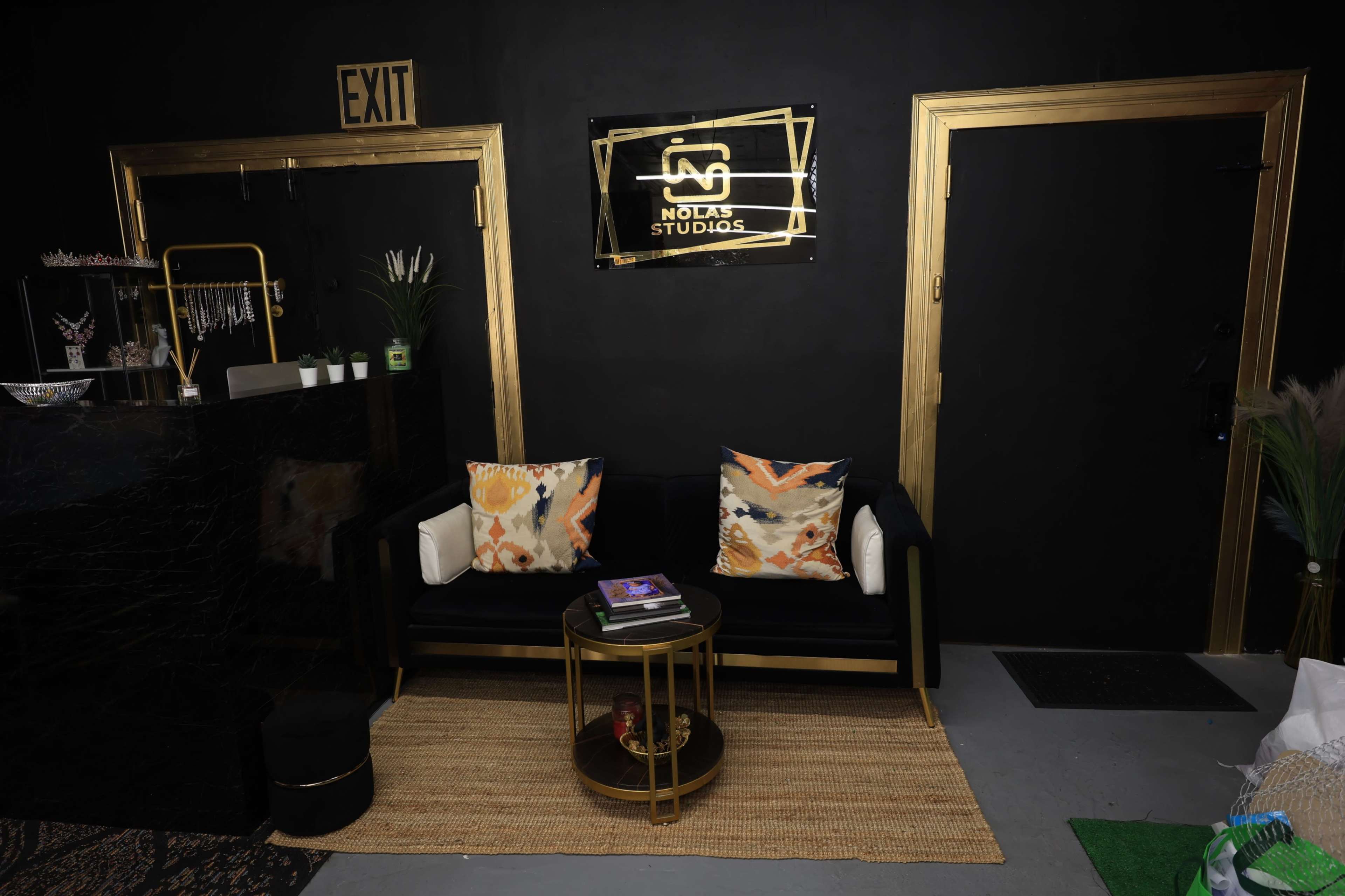 A modern reception area with a black wall, a gold-framed sign reading "Nola's Studios," a black sofa with decorative pillows, and a circular coffee table on a woven mat.