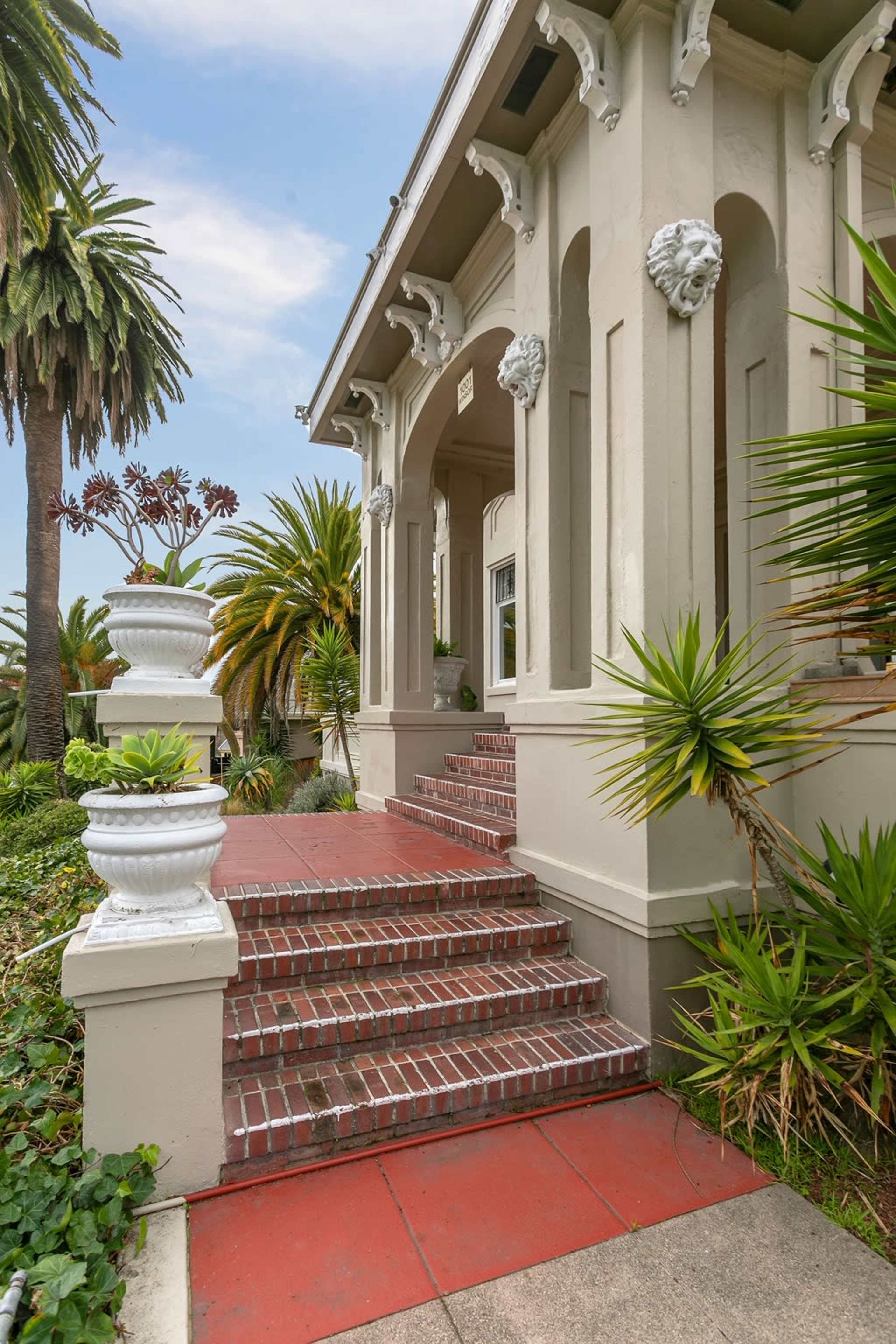 A stately house features a front staircase with brick steps, flanked by white planters and surrounded by palm trees and lush plants.
