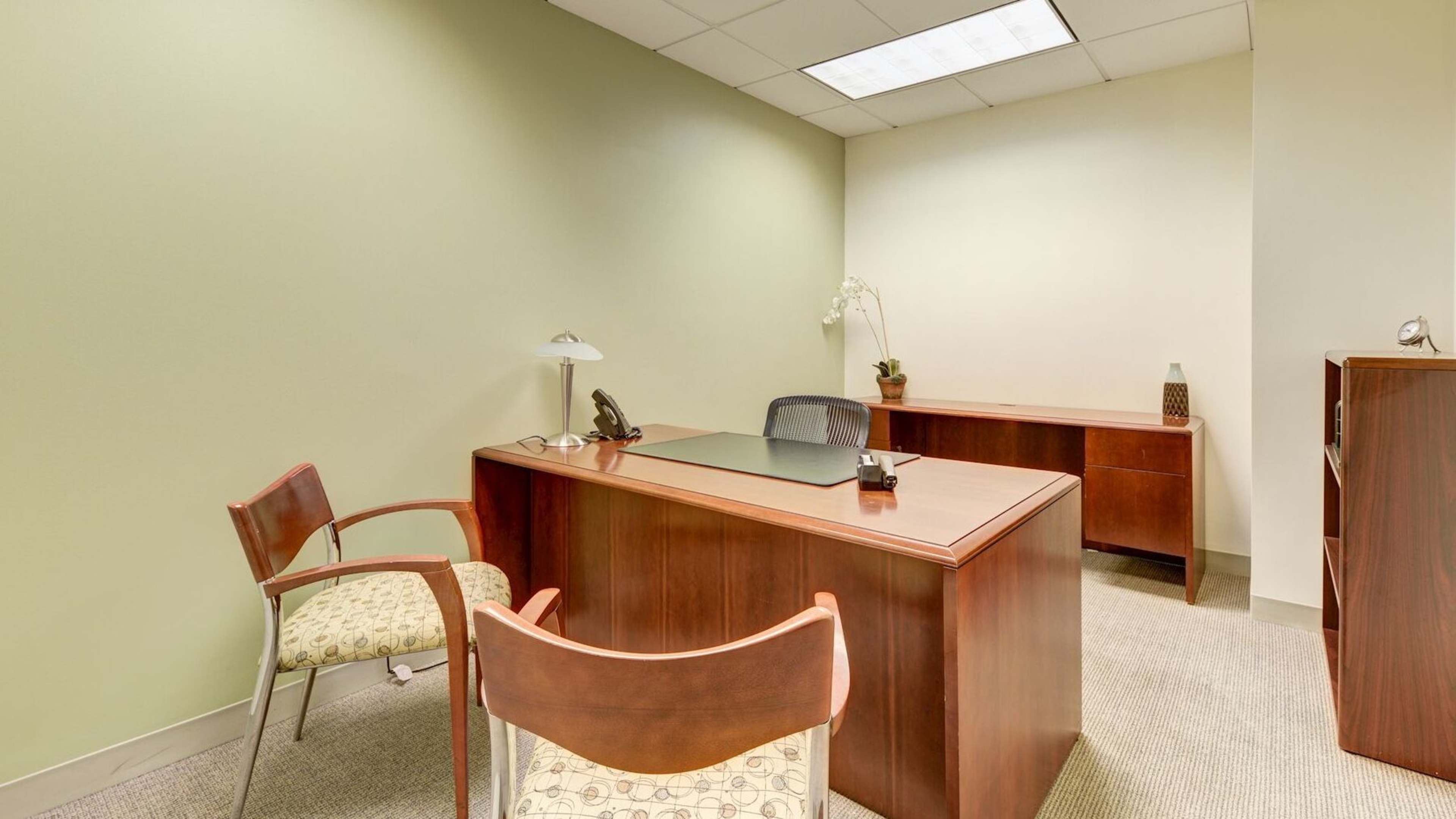 A tidy office space featuring a wooden desk, two chairs, a lamp, and a wall-mounted shelf in a room with green walls.