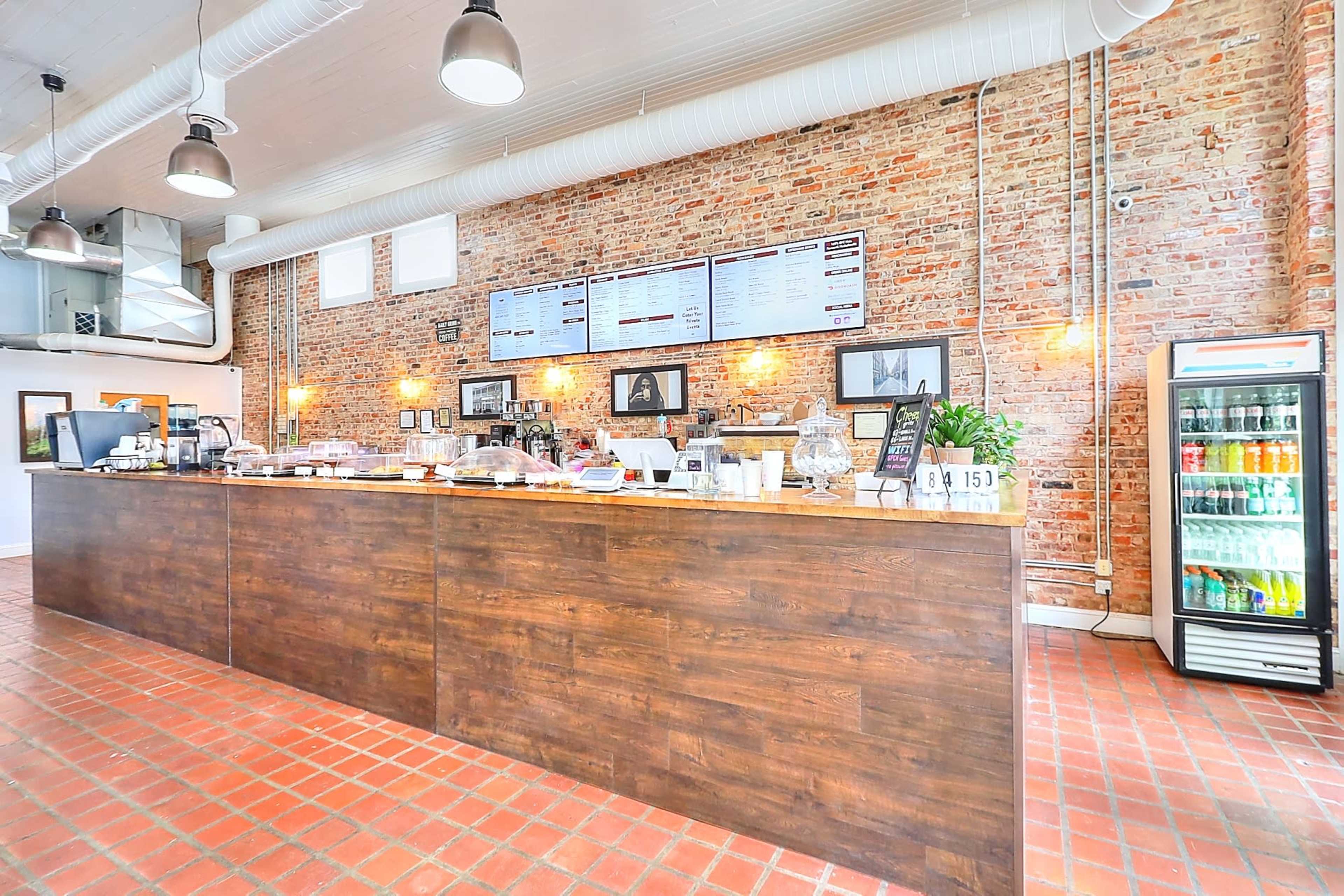 A rustic café interior with a wooden counter, exposed brick walls, and a display of beverages in a glass refrigerator.