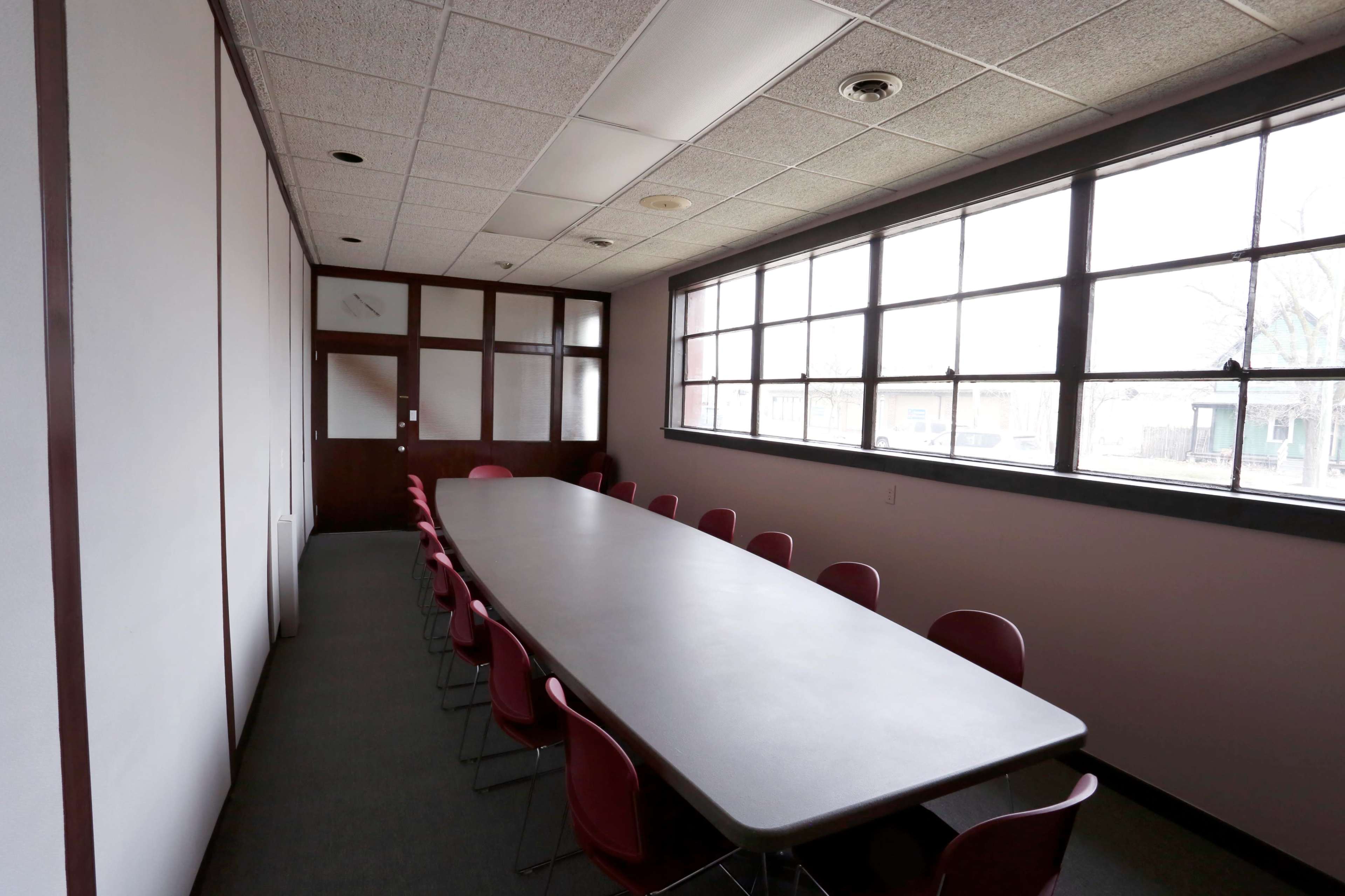 A long table with red chairs is set in a conference room featuring large windows along one wall.