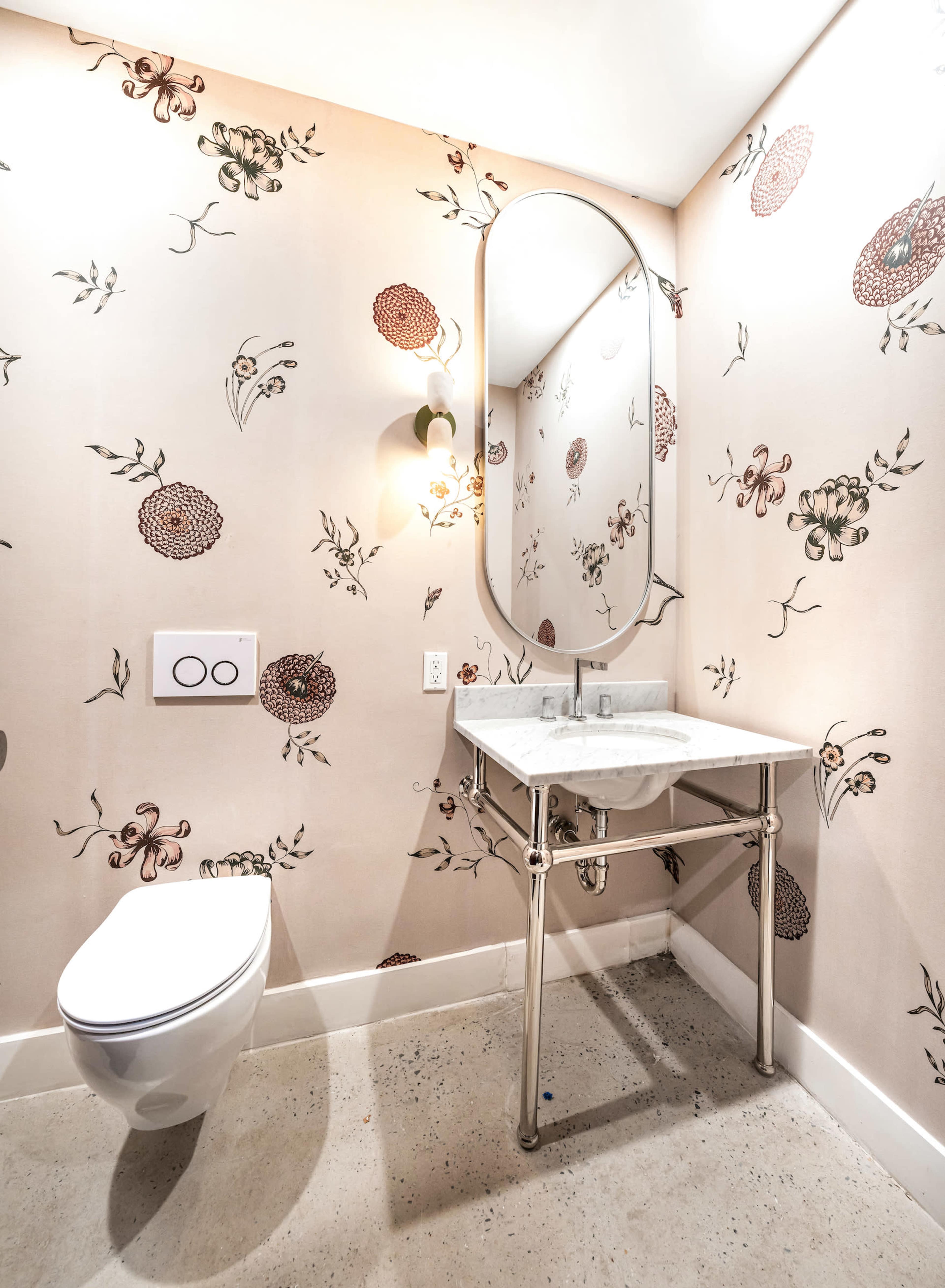 The image shows a small bathroom featuring floral wallpaper, a wall-mounted toilet, and a marble-topped vanity with a round mirror.
