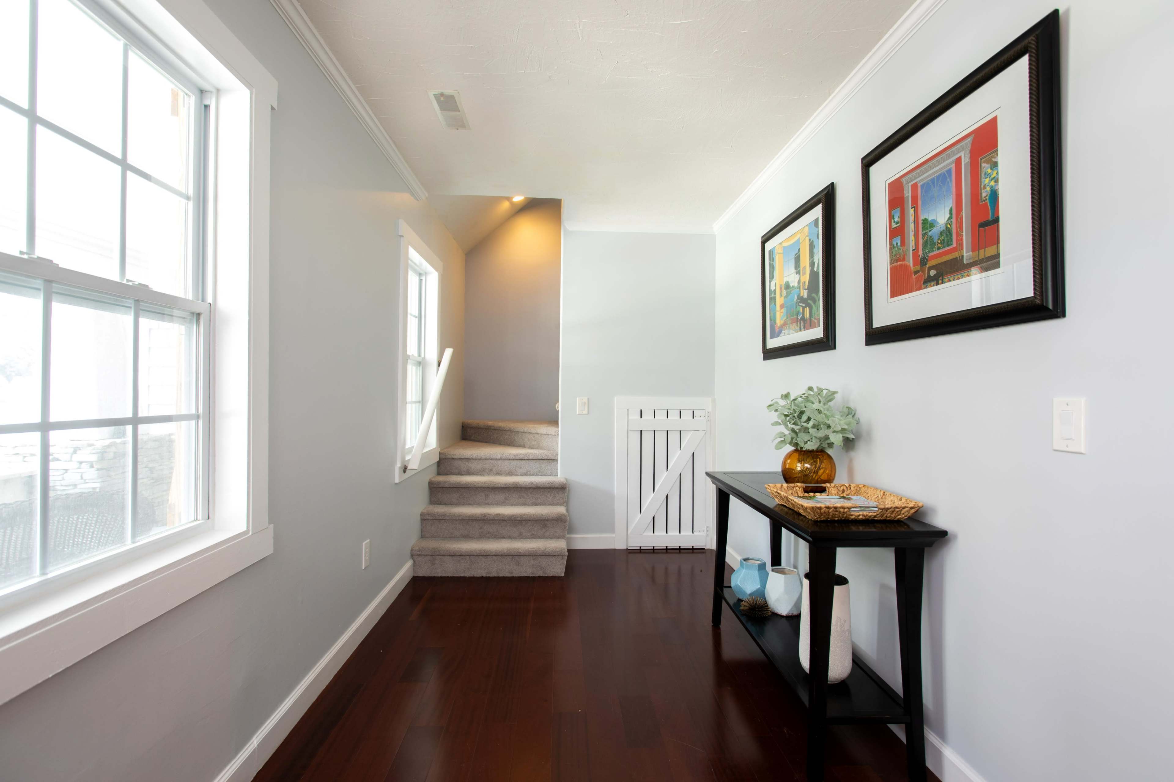 A well-lit hallway with a staircase at the end features two framed artworks on the wall and a console table with decorative items.