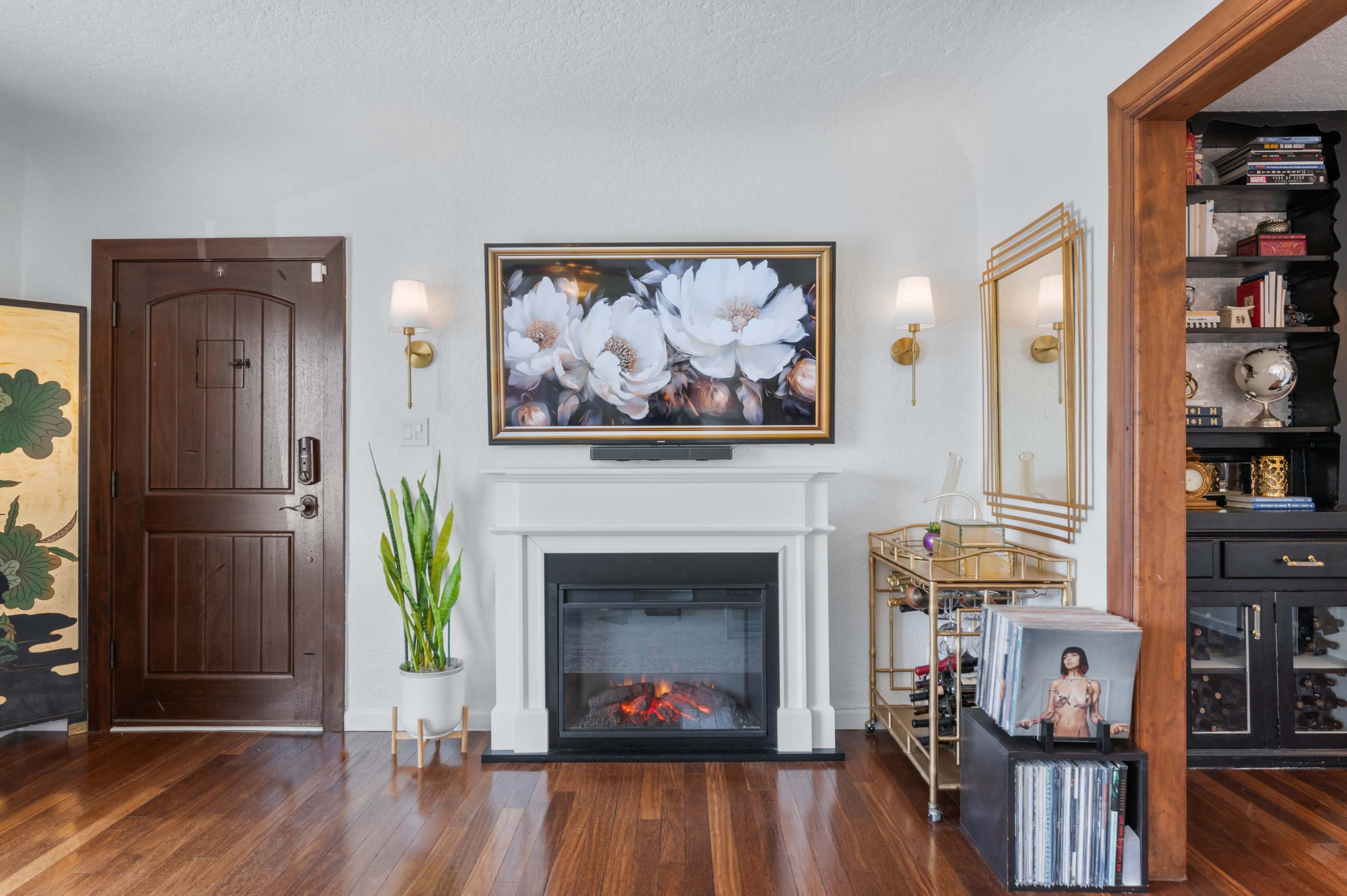 A living room featuring a fireplace with a mounted television displaying flower artwork, a wooden door, and shelves with various decorative items.