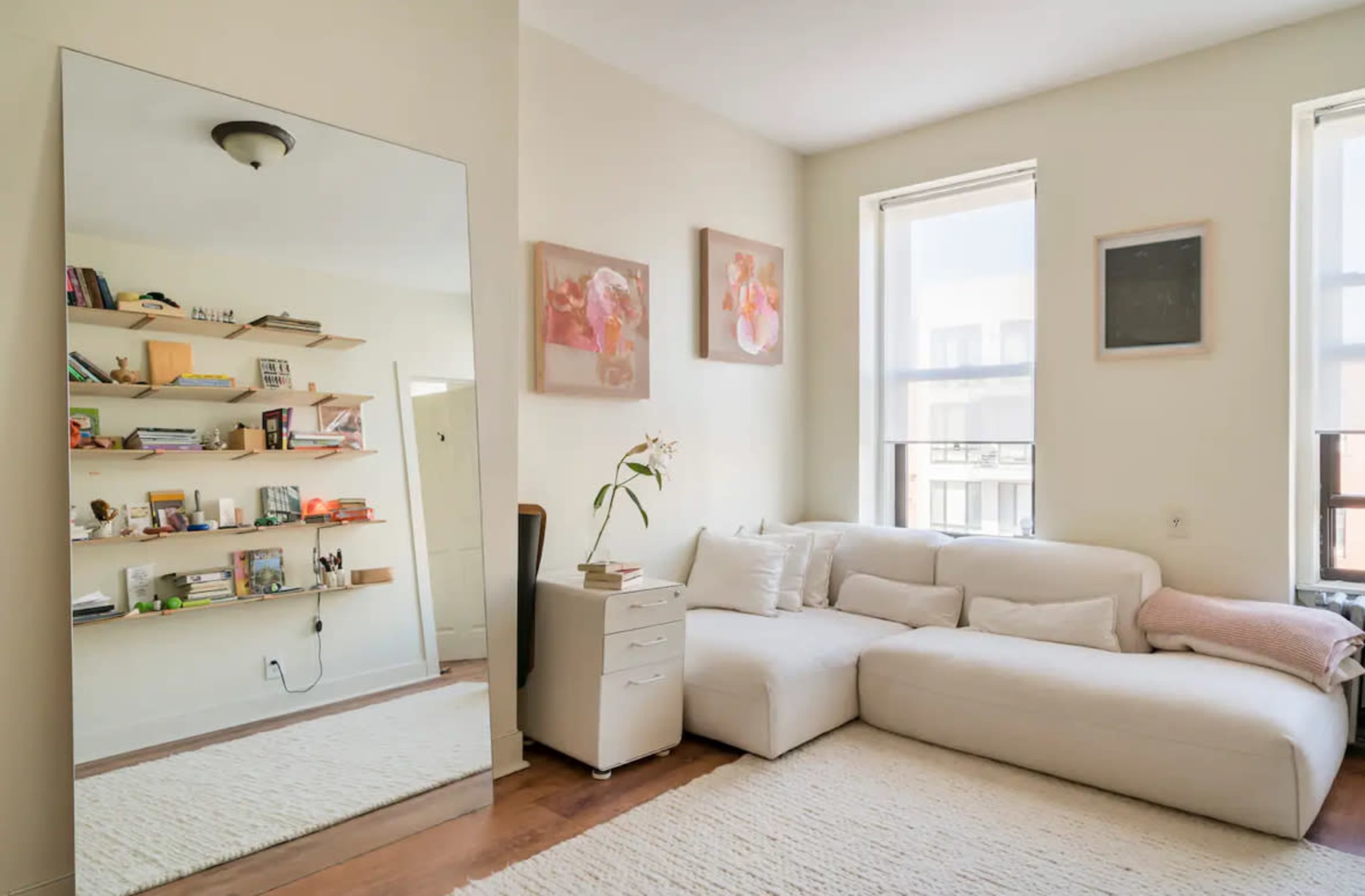 The image shows a bright living room featuring a large mirror, a white sectional sofa, and a shelf with books and decorative items.
