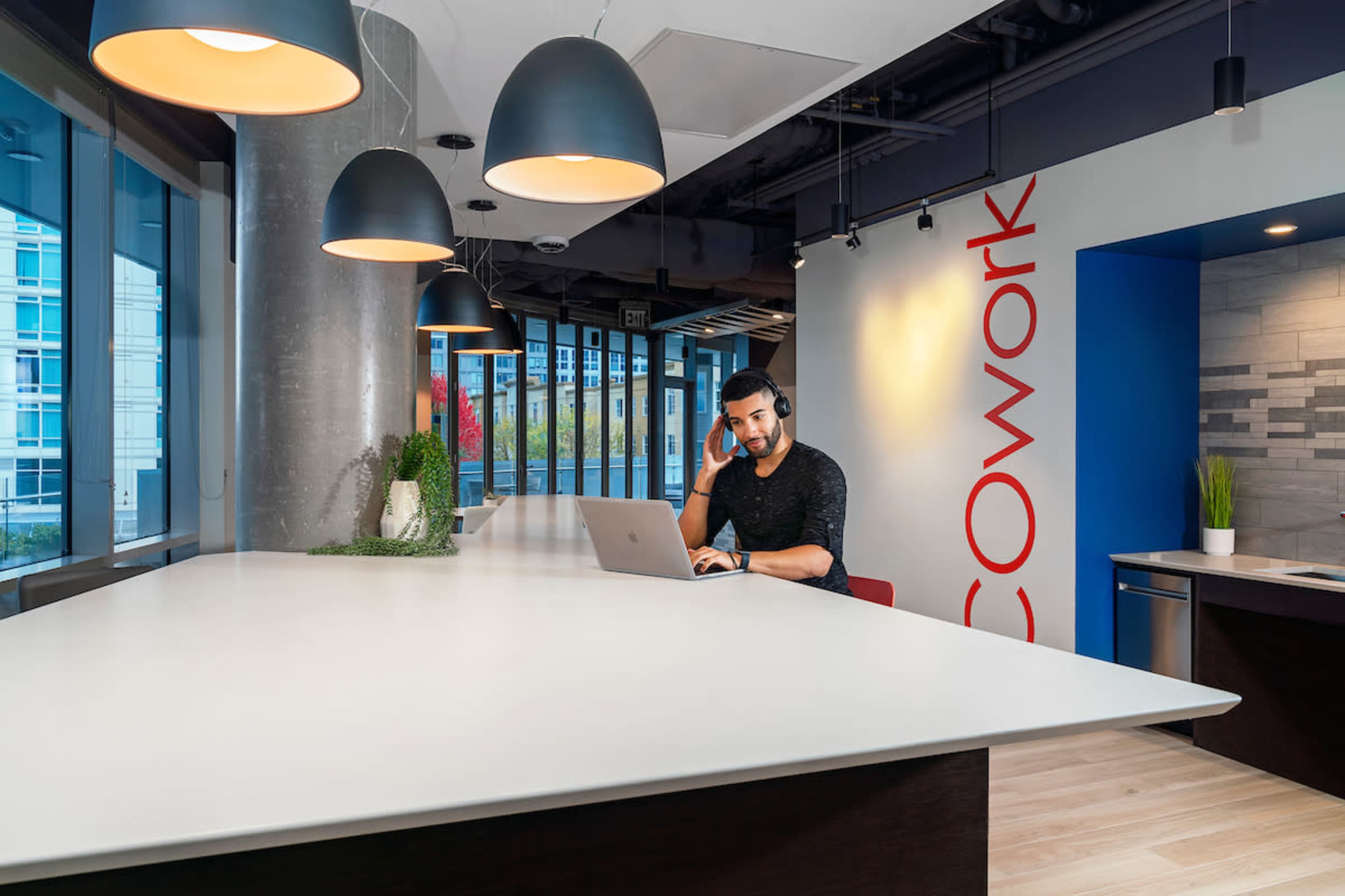 A man is sitting at a large, white table working on a laptop while holding a phone to his ear in a modern office space.