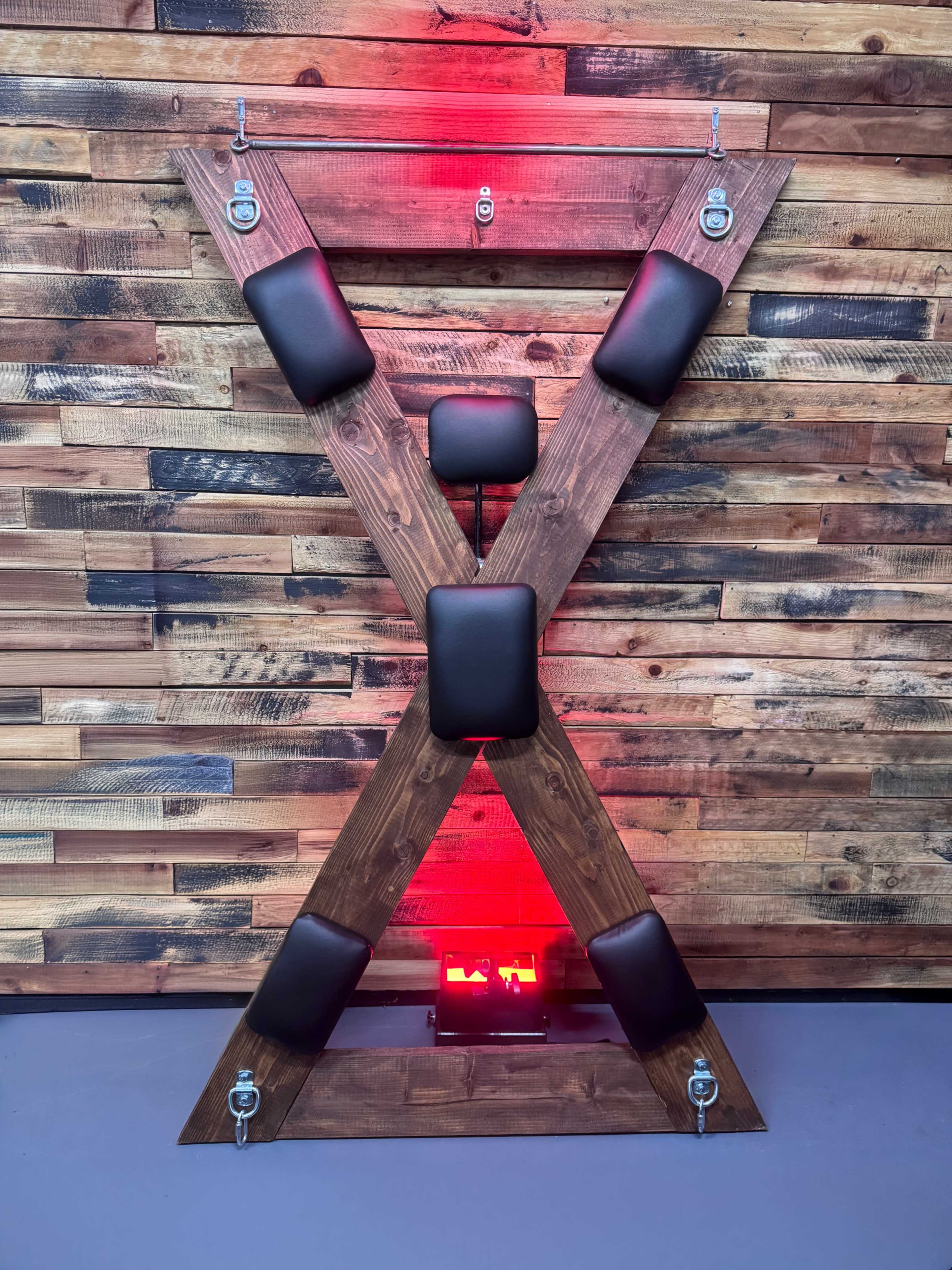 The image features an X-shaped wooden structure with padded black cushions attached, set against a backdrop of wooden planks illuminated by red lighting.