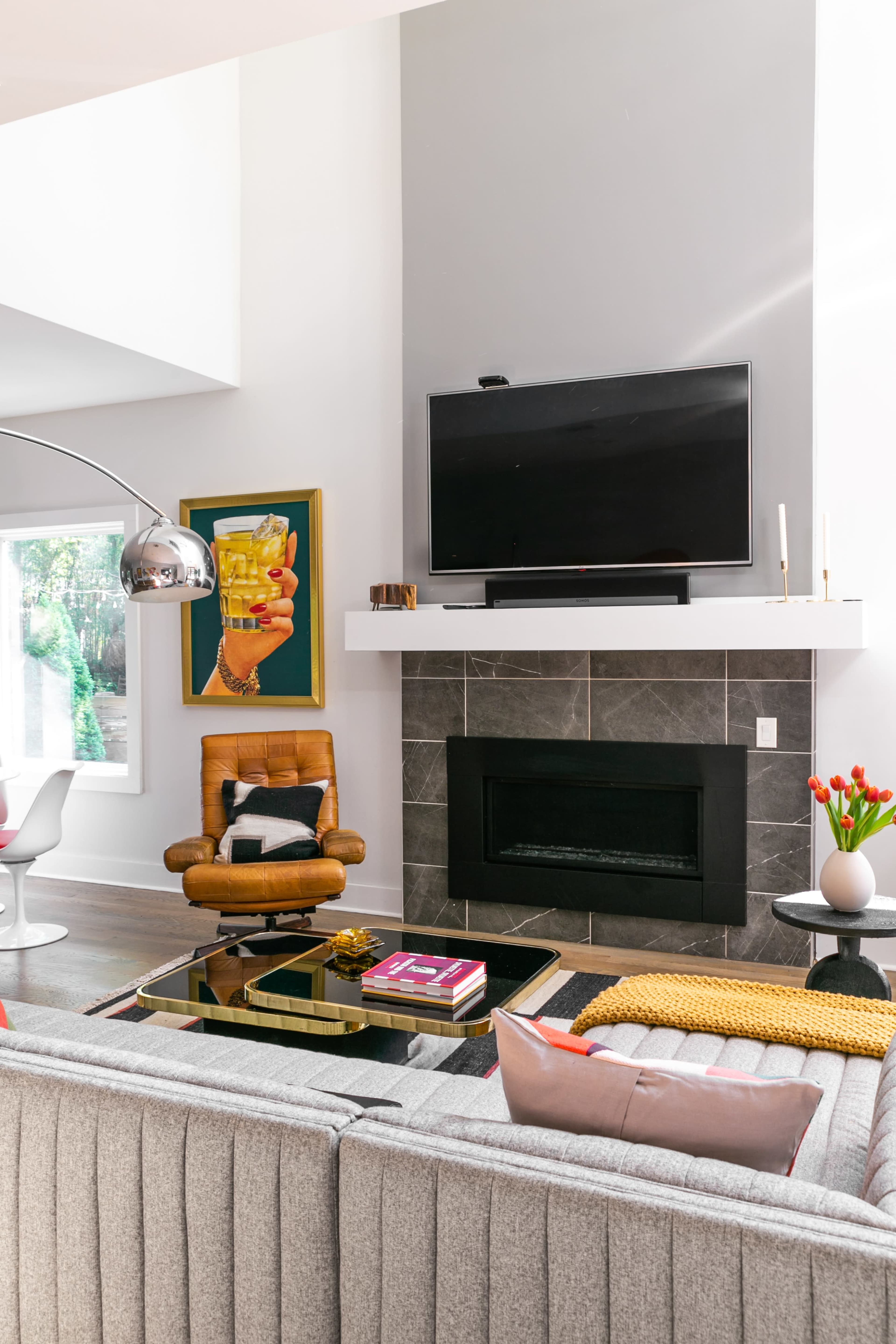 A modern living room features a gray sectional sofa, a yellow throw rug, a flat-screen TV above a stone fireplace, and a mid-century armchair, with a vibrant artwork and decorative items enhancing the space.
