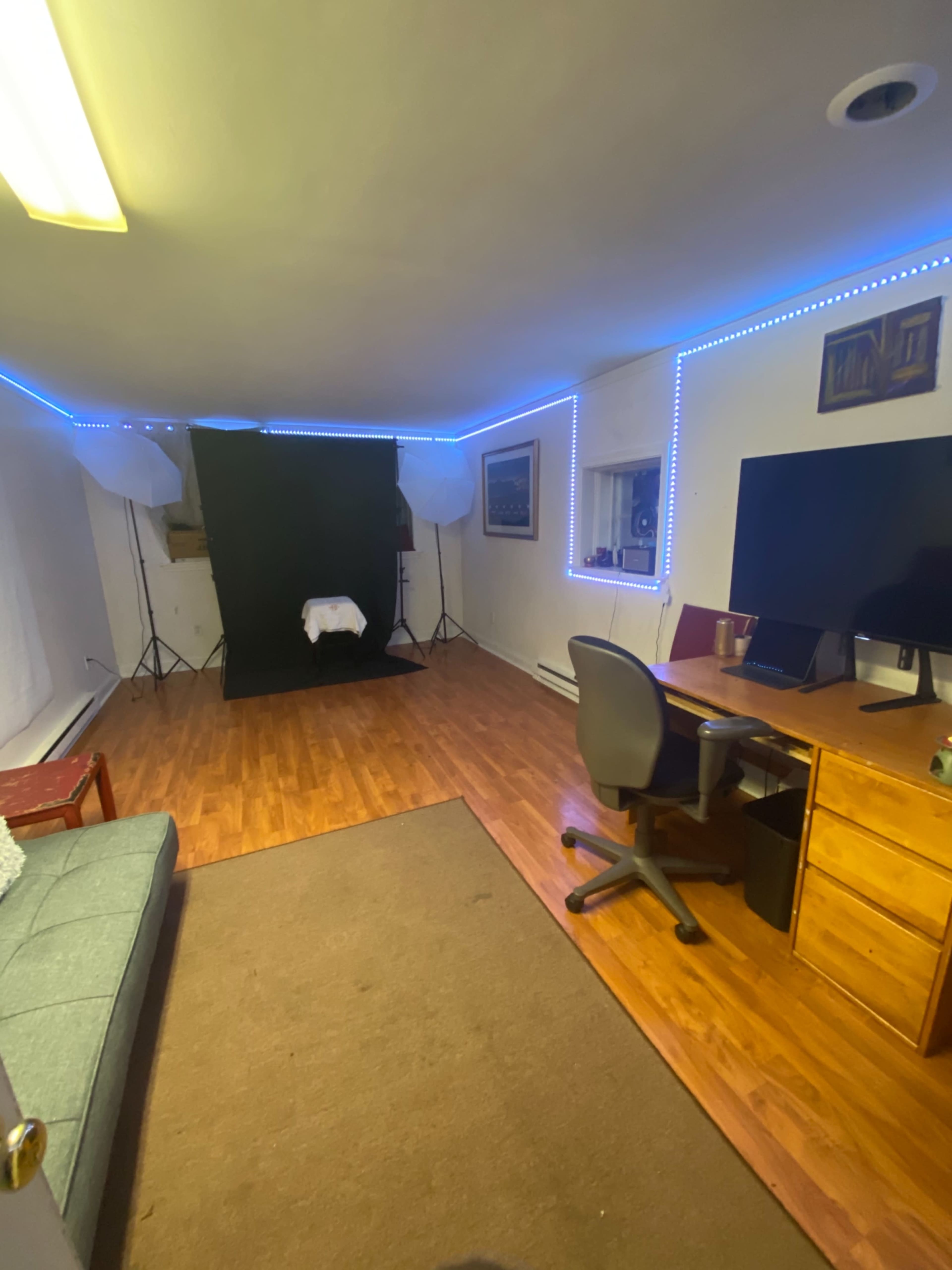 The image shows a well-lit room with wooden flooring, featuring a sofa, a desk with a computer, and a backdrop setup for photography.