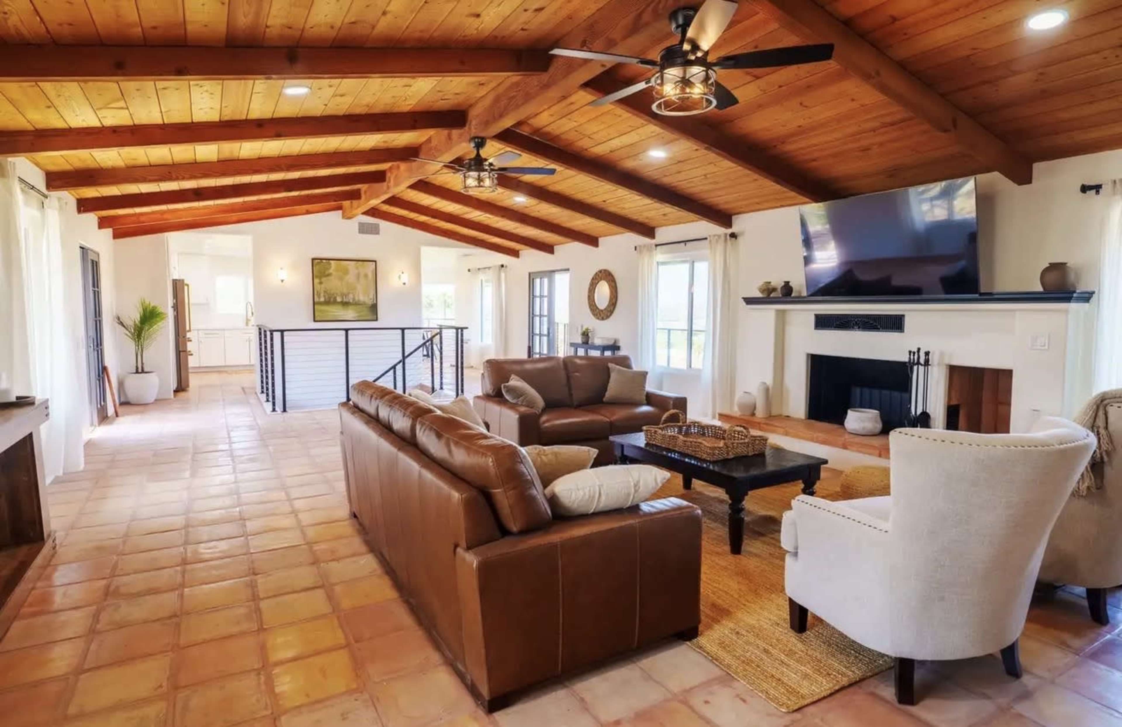 The image shows a spacious living room with a high, wooden ceiling, featuring a seating area with leather sofas, a coffee table, and a fireplace.