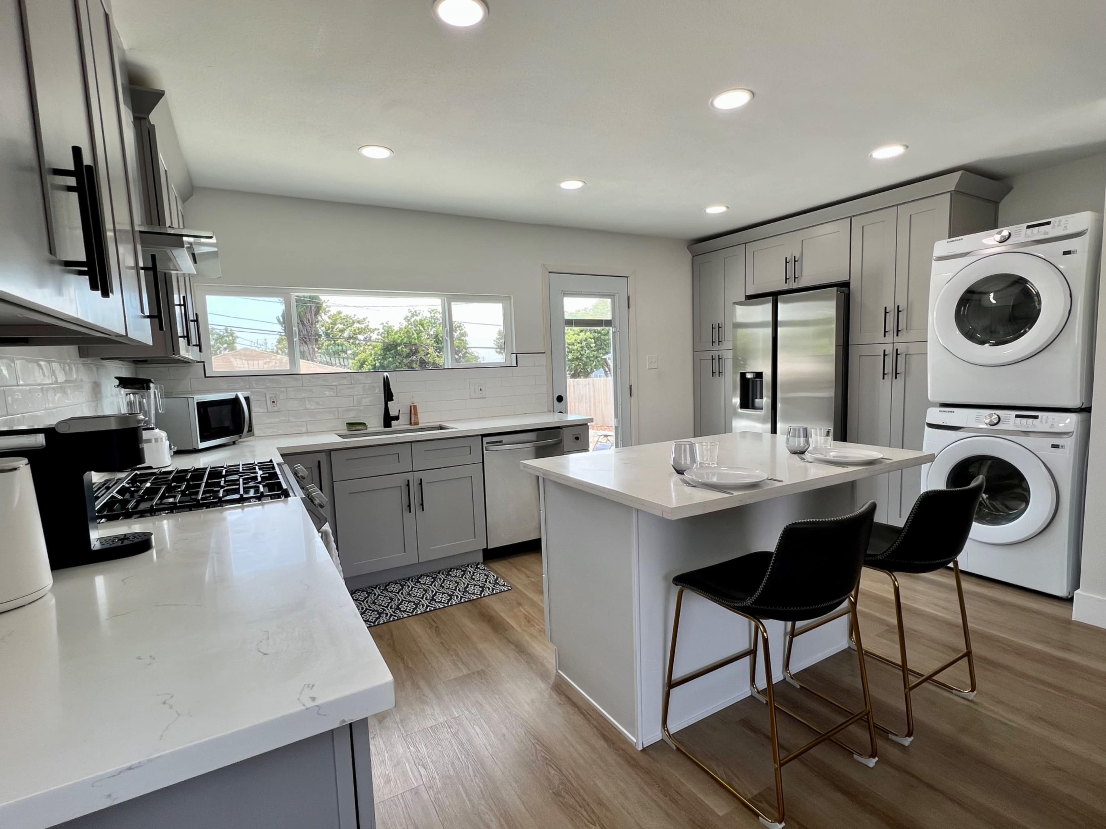 A modern kitchen featuring gray cabinets, a central island with seating, and a stacked washer and dryer unit.